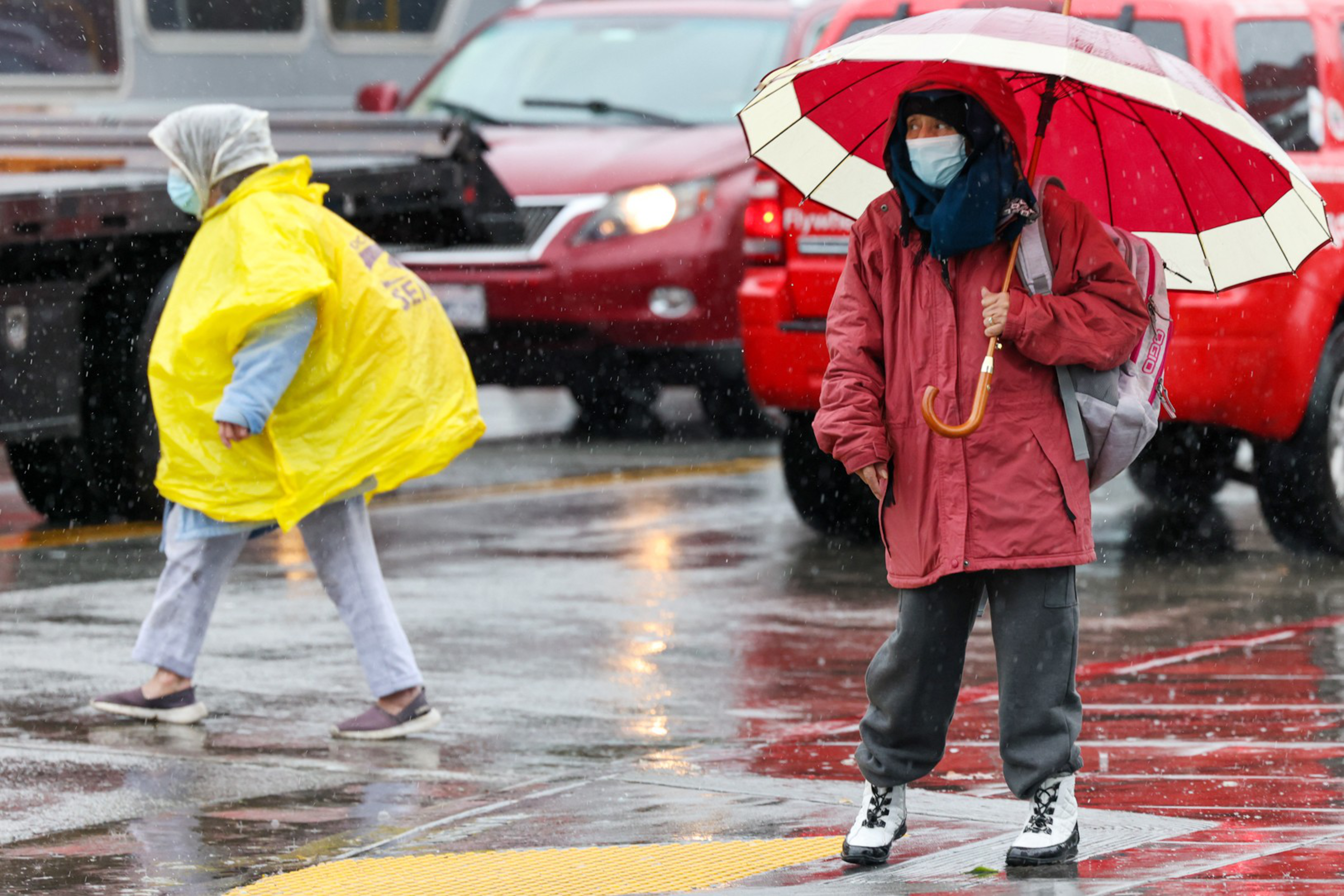 A person in a red coat and face mask holds a red-and-white umbrella, while another in a yellow rain poncho walks across a wet street with cars in the background.