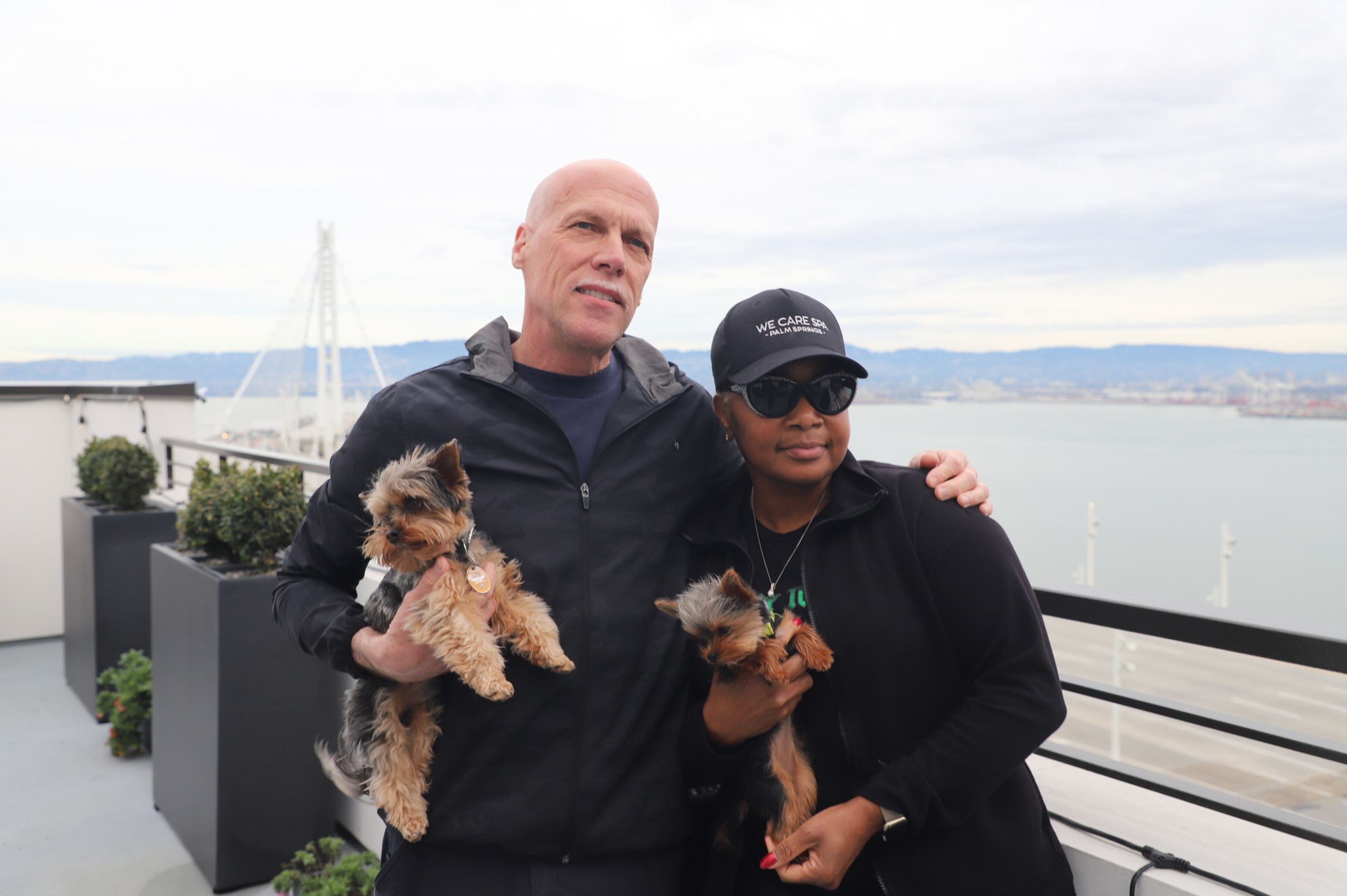 A couple stands on a balcony, holding their two small dogs.