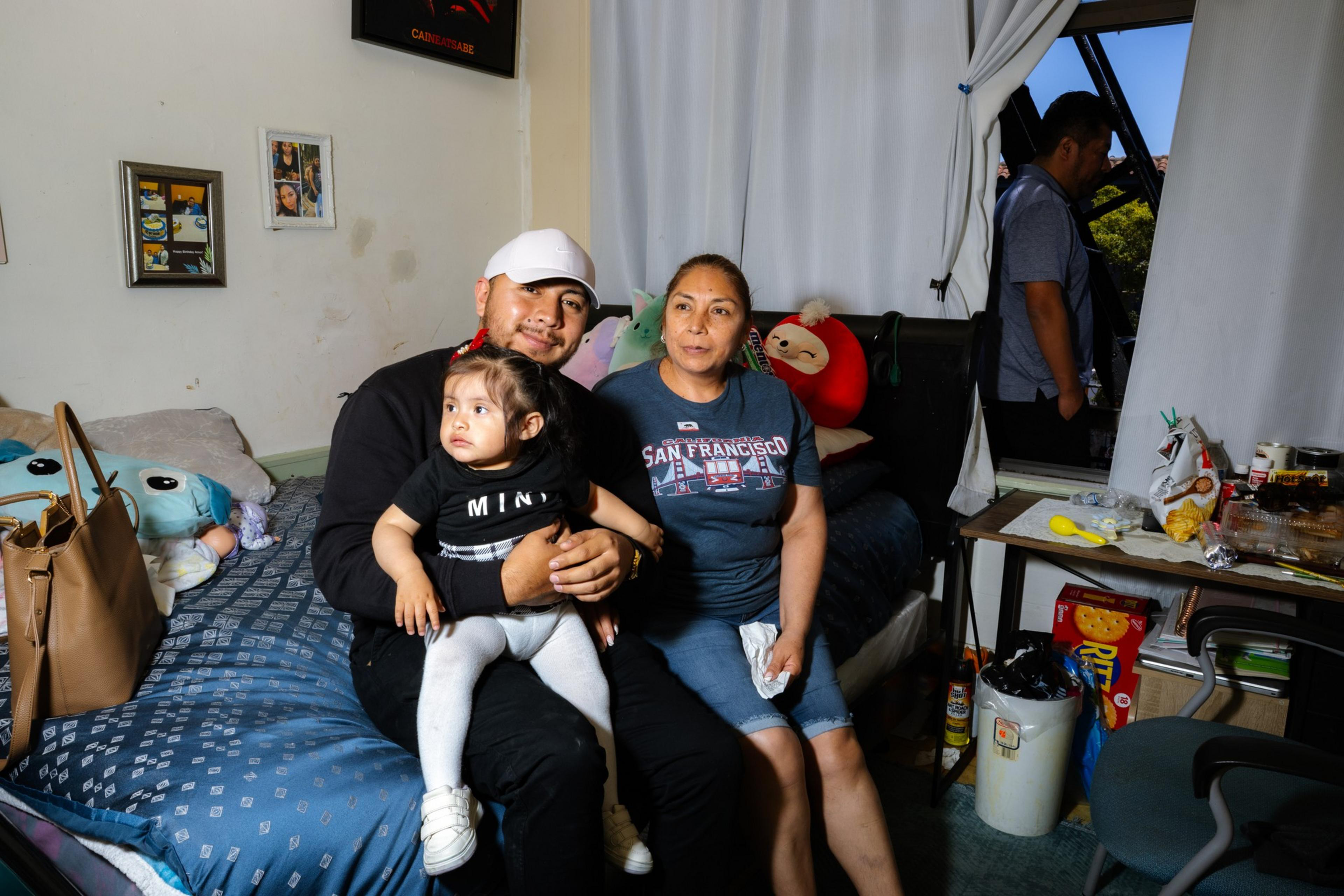 A family of three poses on a bed with a child sitting on an adult's lap. There's a stuffed toy behind them, a cluttered table, and a person near the window.