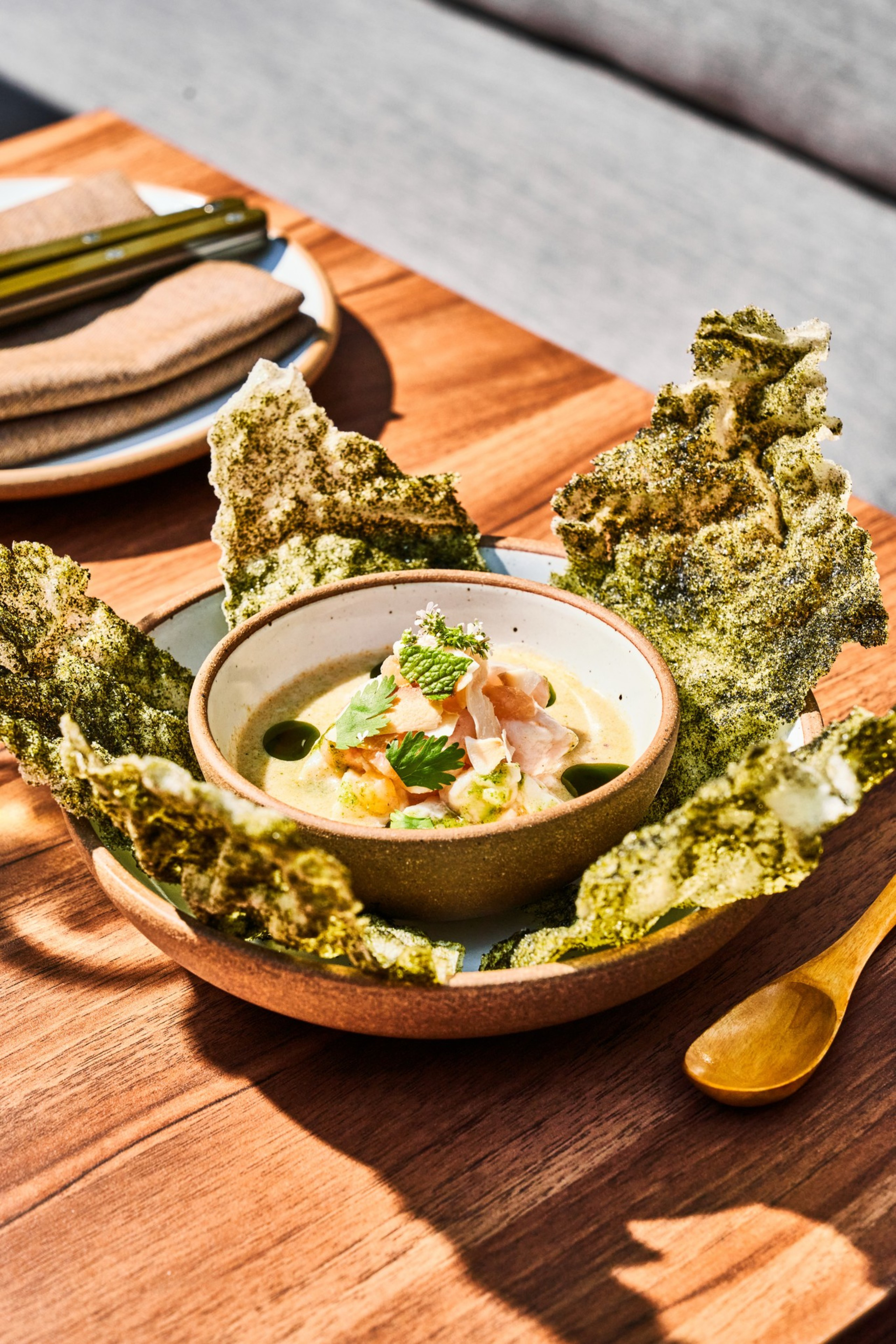 A ceramic bowl holds a creamy dish topped with herbs, surrounded by large, crispy seaweed on a wooden table, next to a wooden spoon and folded napkins on a plate.