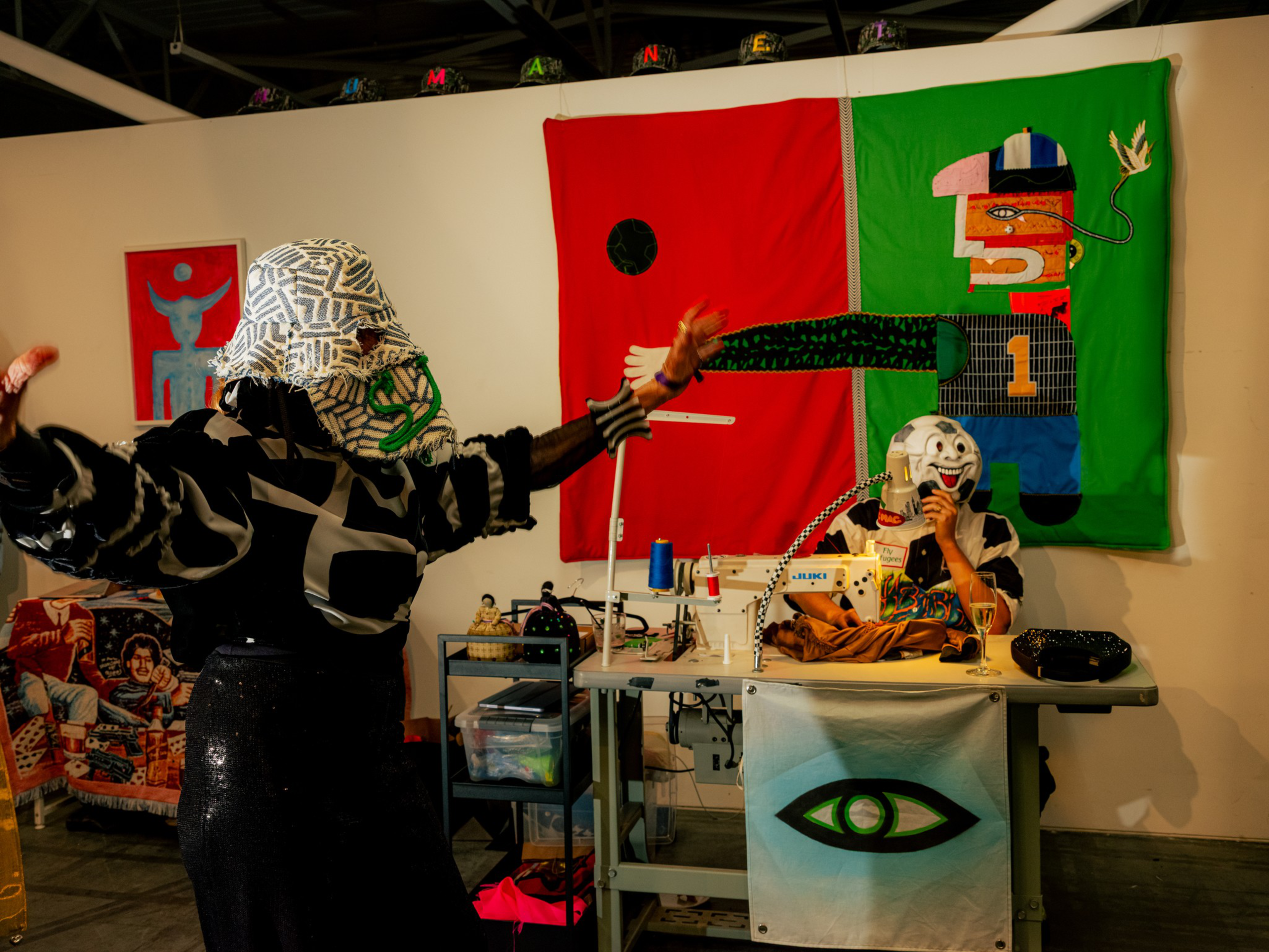 Two people wearing patterned masks and black-and-white clothing interact in a colorful room with a sewing machine and abstract art on the walls.