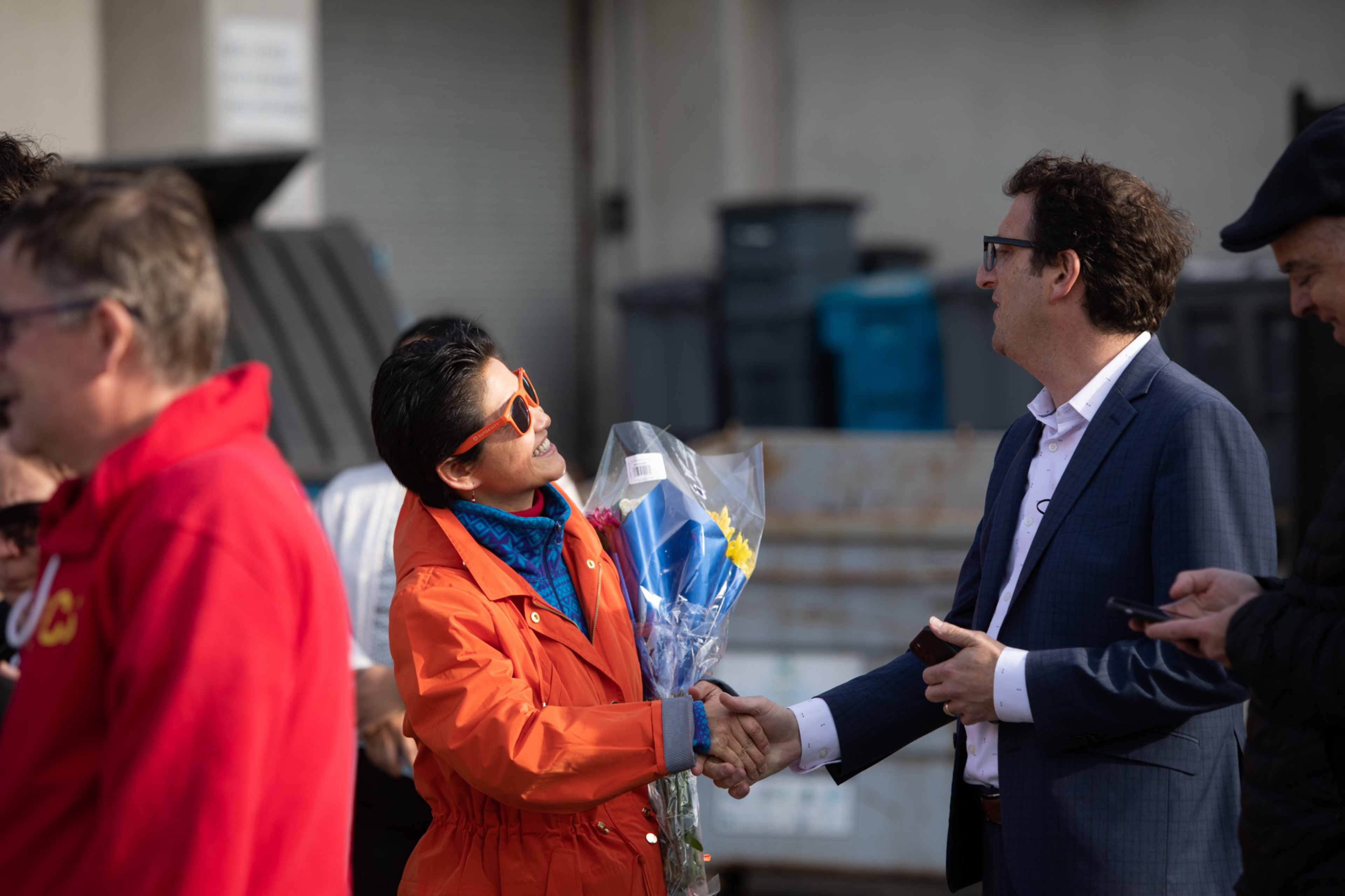 A person in an orange jacket and sunglasses shakes hands with a man in a suit while holding a bouquet of flowers wrapped in plastic.