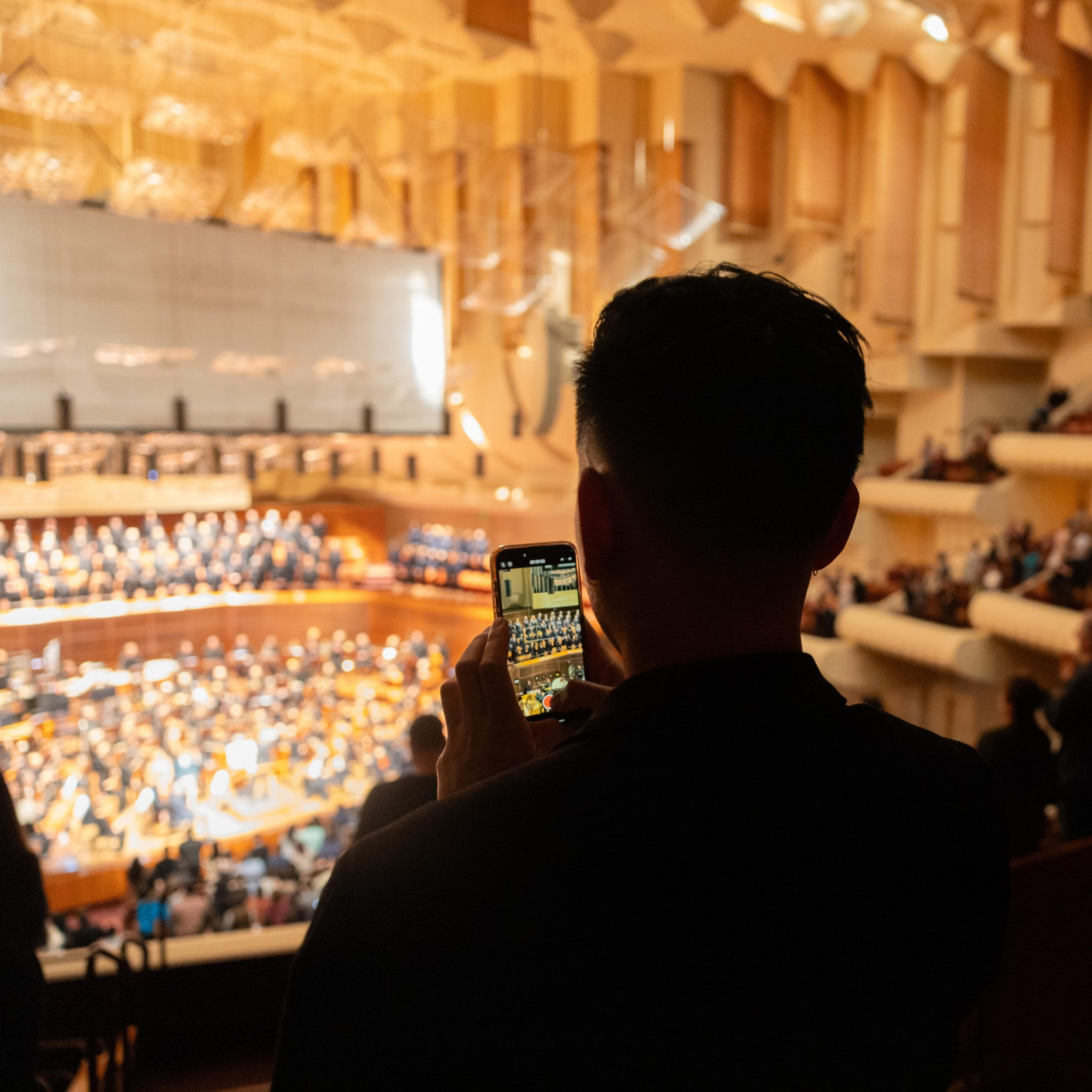 A person is taking a photo of an orchestra in a concert hall with their smartphone.