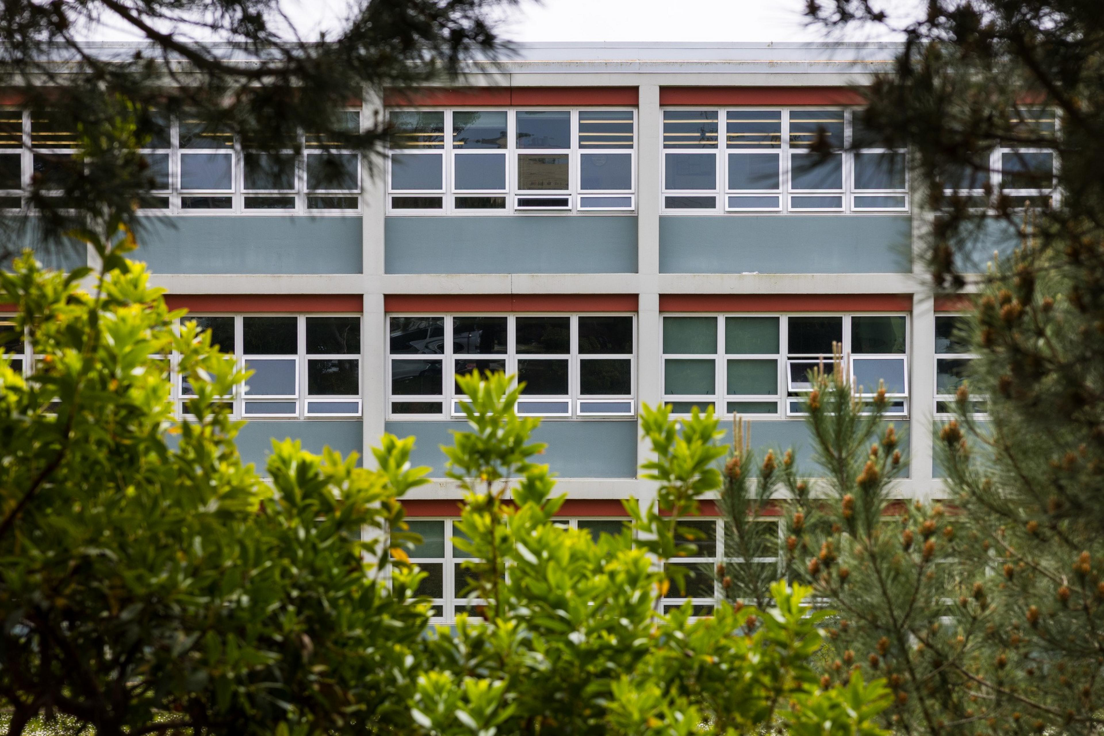 The image shows a multi-story building with large rectangular windows, framed by leafy green trees in the foreground, suggesting a school or office.