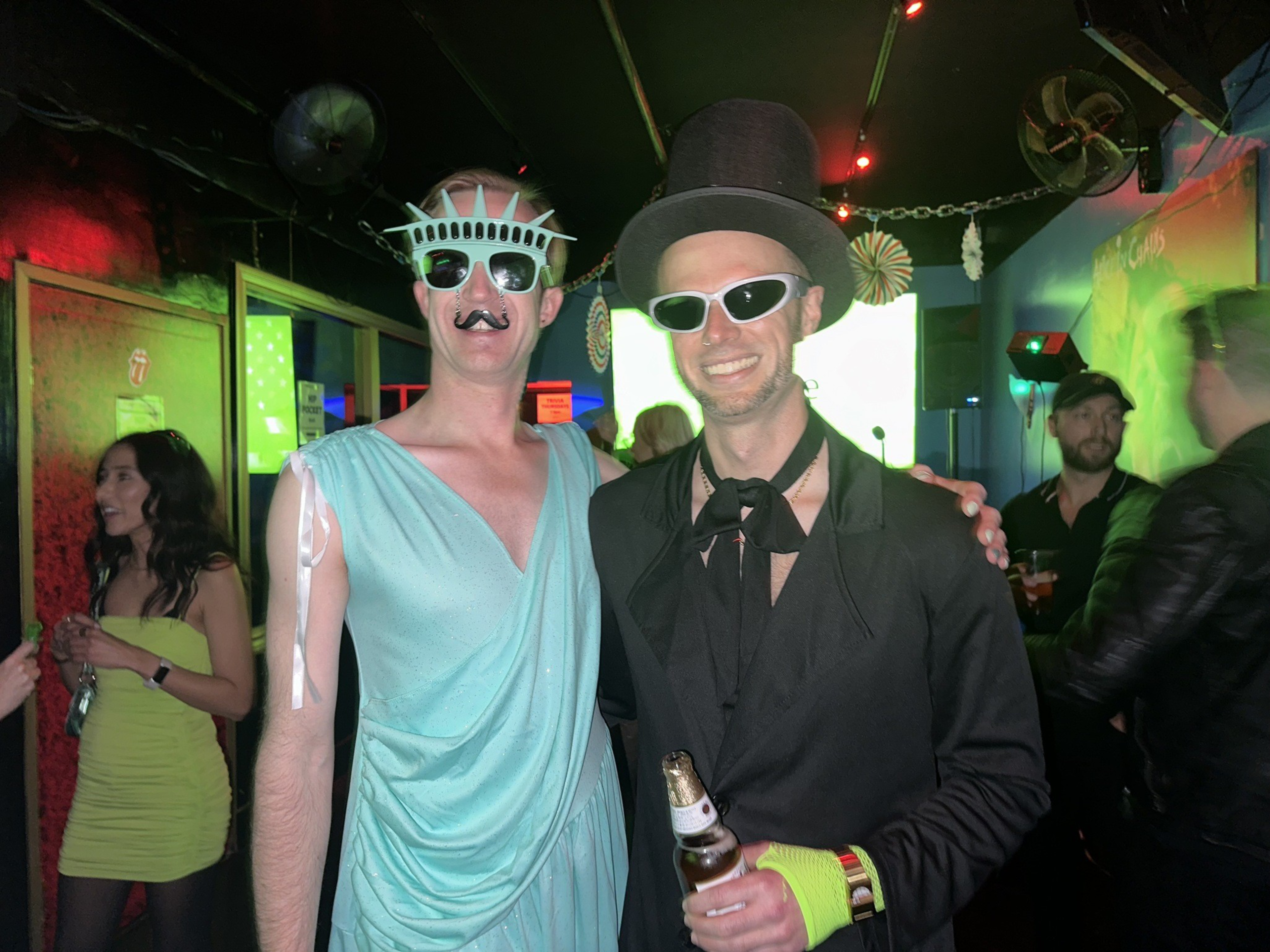 Two people in costumes pose together; one wears a Statue of Liberty outfit with sunglasses and a mustache, the other in a black suit with a top hat and sunglasses.