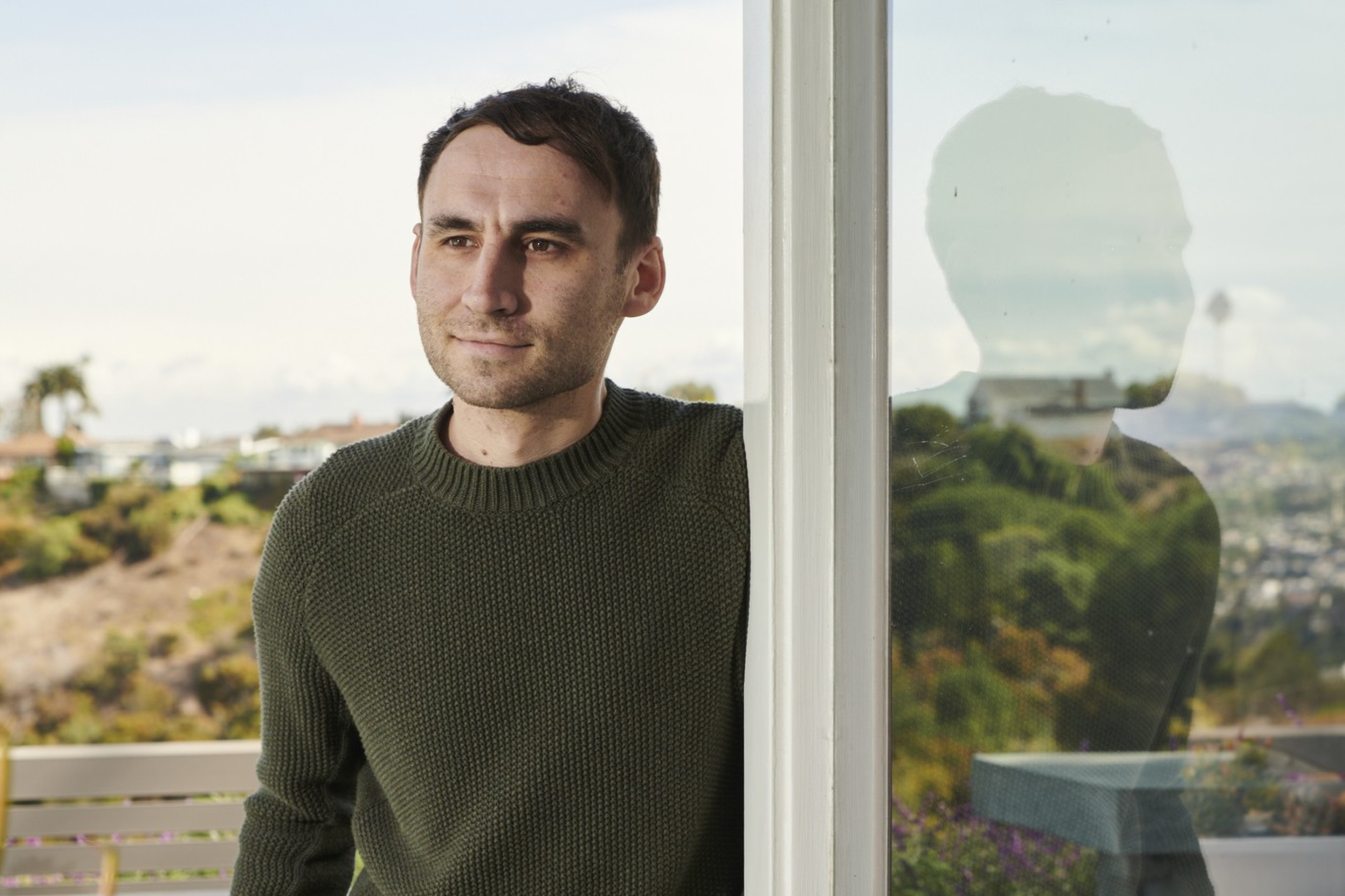 A person in a green sweater stands by a glass wall, hands in pockets. There's a reflection in the glass and a blurred outdoor view with plants and a clear sky.
