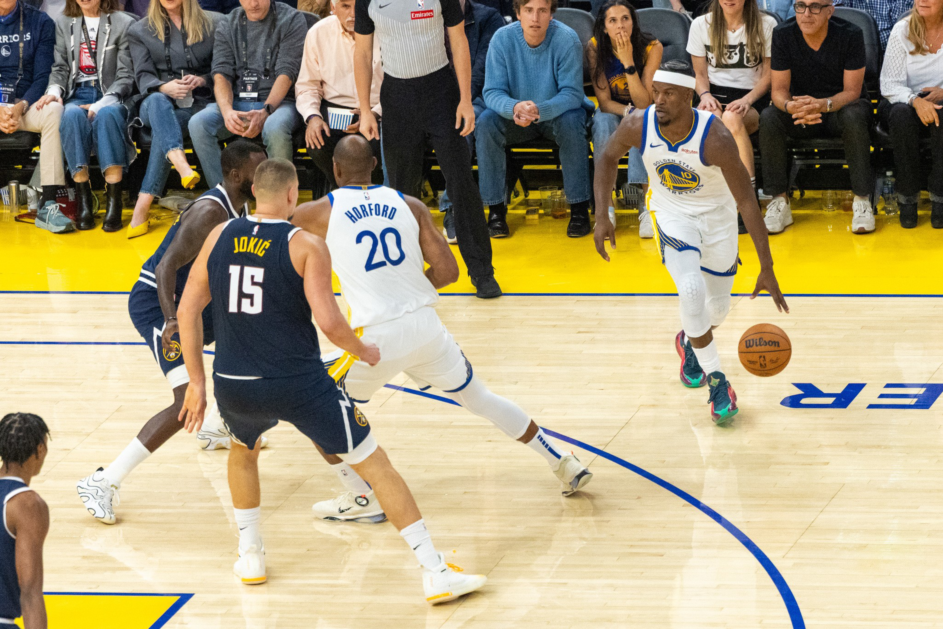 A basketball player in a white Golden State Warriors uniform dribbles the ball past defenders from the Denver Nuggets near the three-point line.