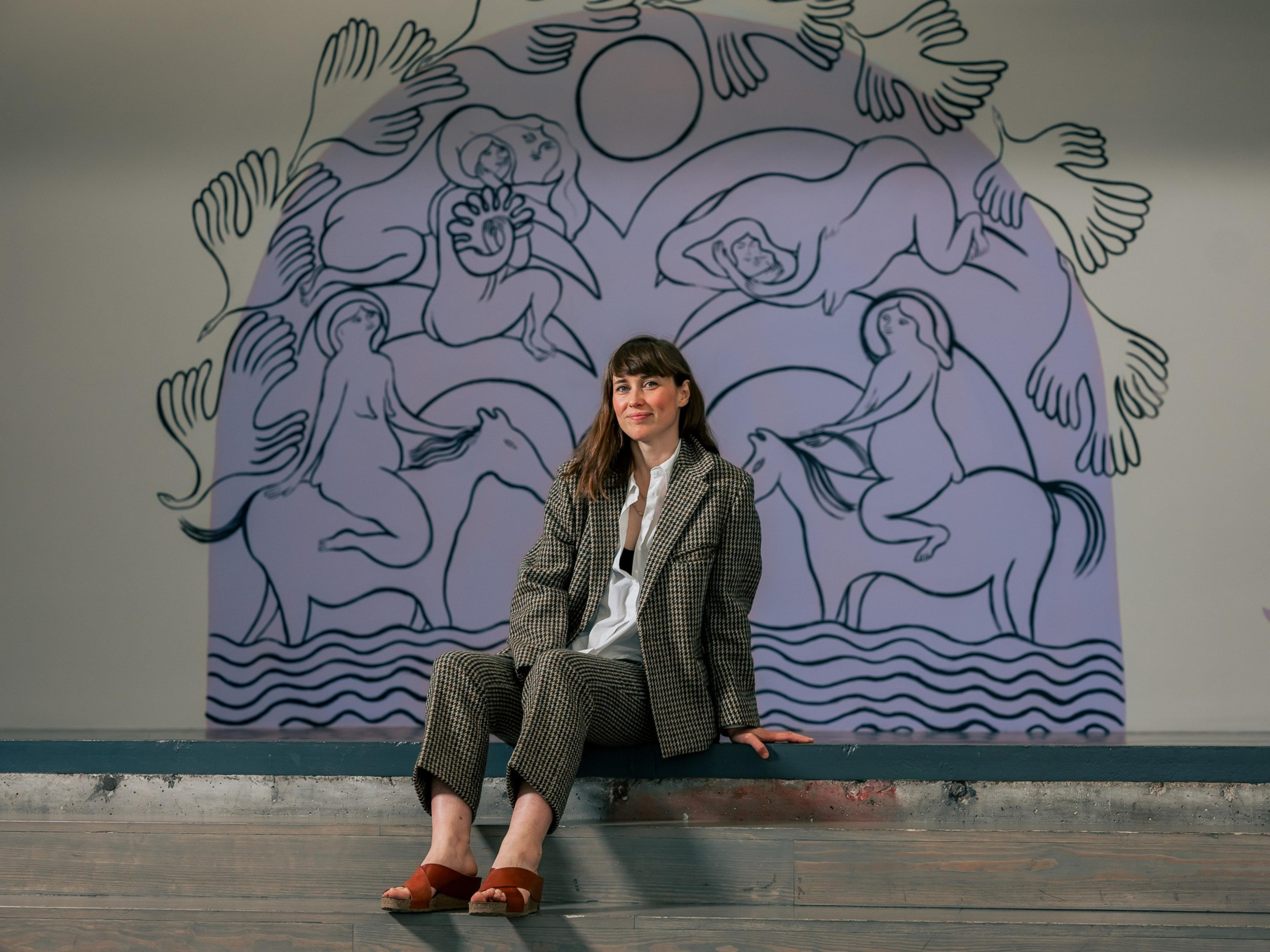 A woman in a checkered suit sits on a ledge in front of a large mural. The mural depicts abstract figures and hands in shades of blue and purple.