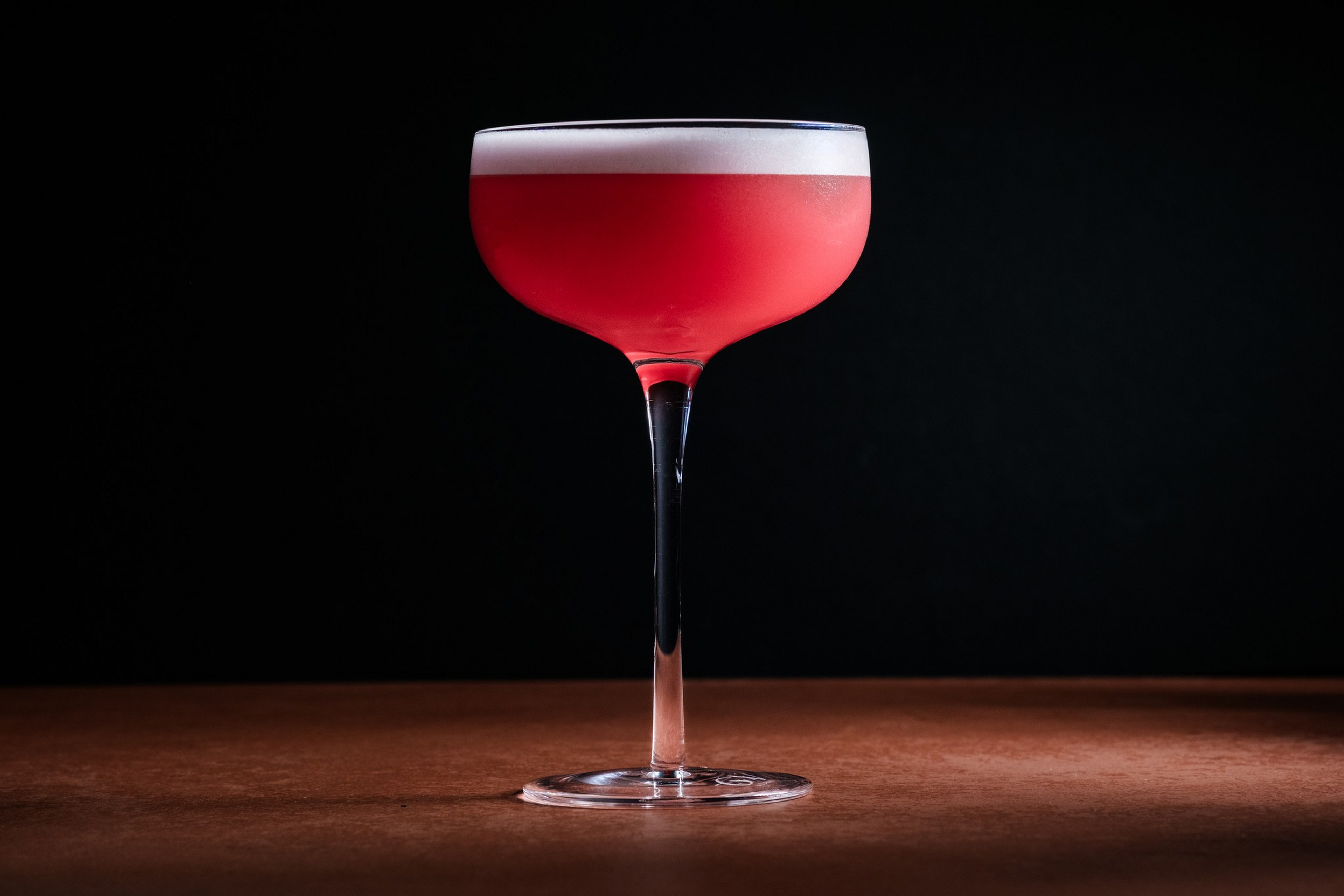 A tall, elegant glass holds a bright pink cocktail with a frothy white foam layer on top, set against a dark background.