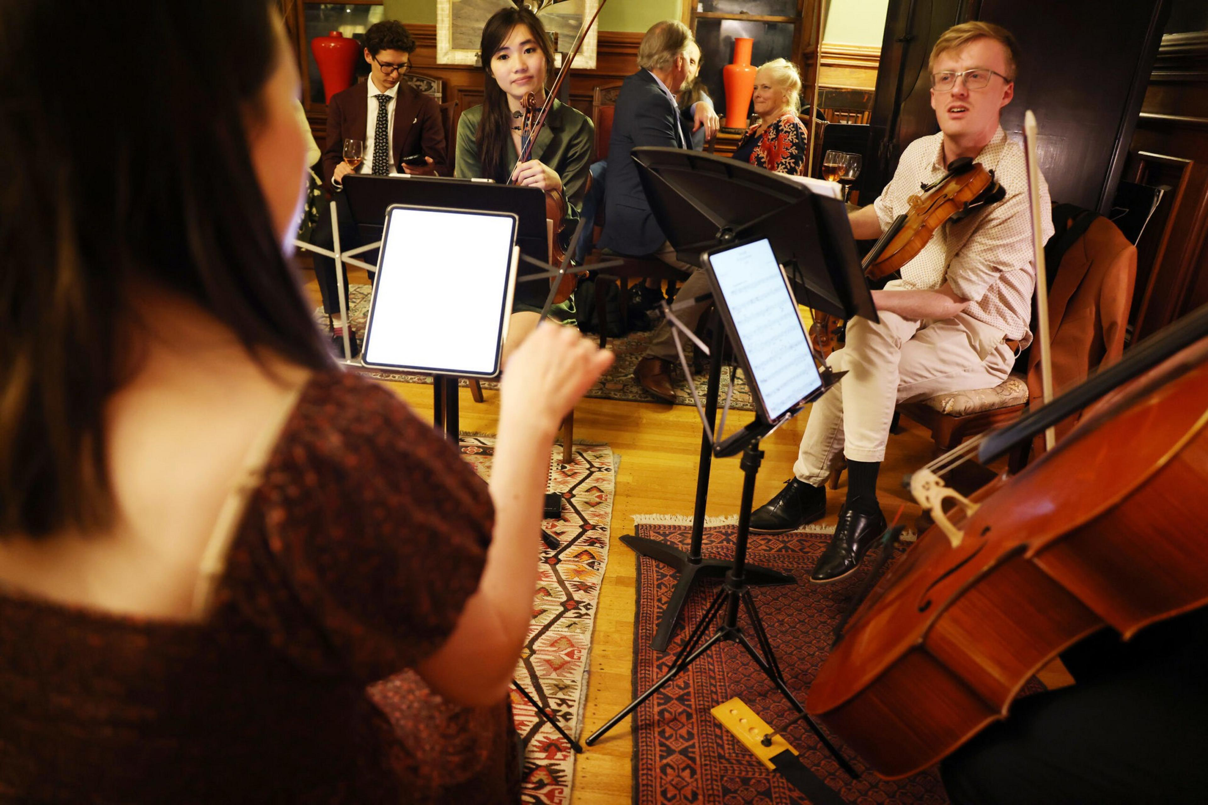 Three musicians play string instruments while reading sheet music as two people converse in the background in a warmly lit, cozy room.