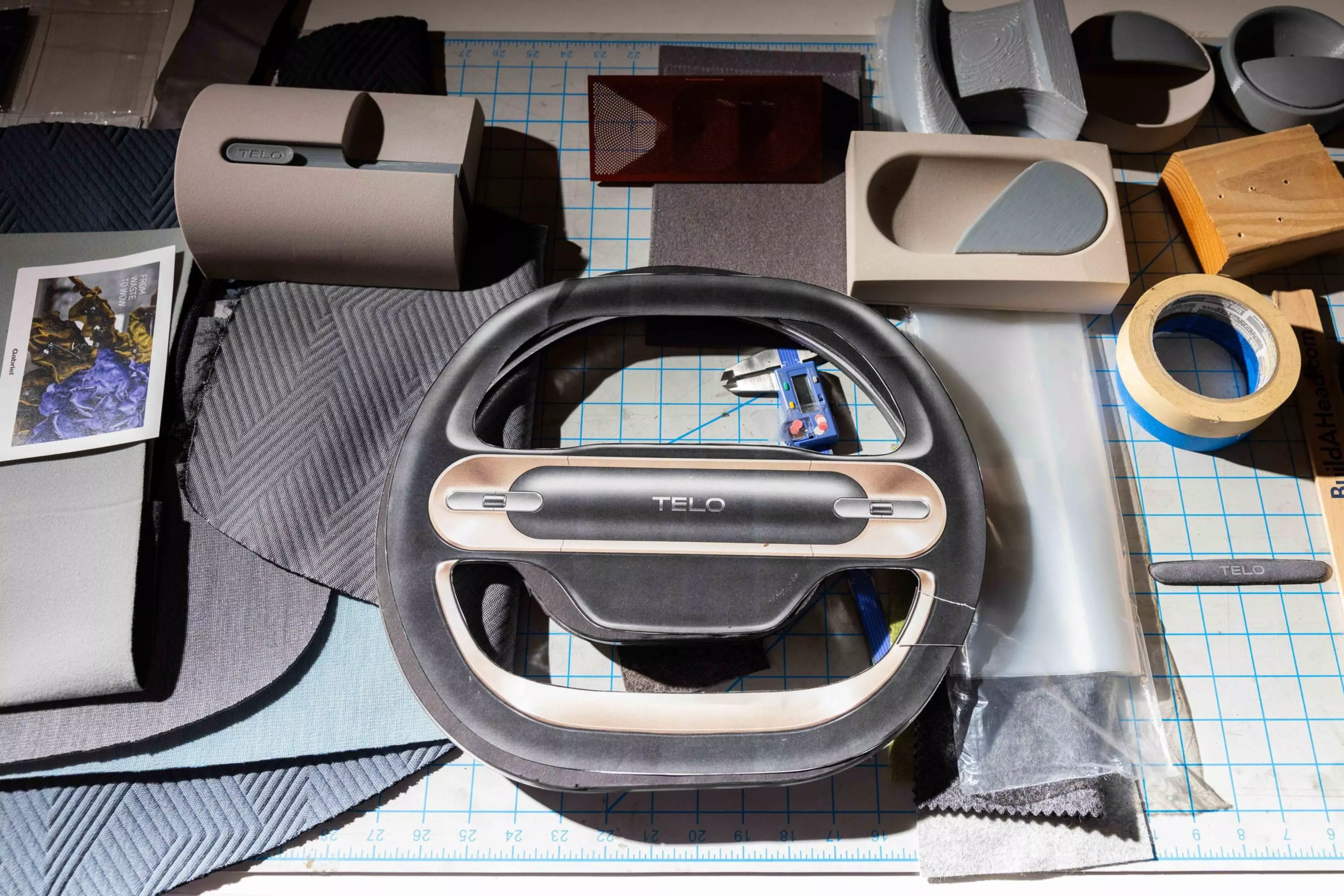 A black and beige TELO steering wheel sits on a grid cutting mat surrounded by various fabric samples, foam pieces, a caliper, masking tape, and a card photo.
