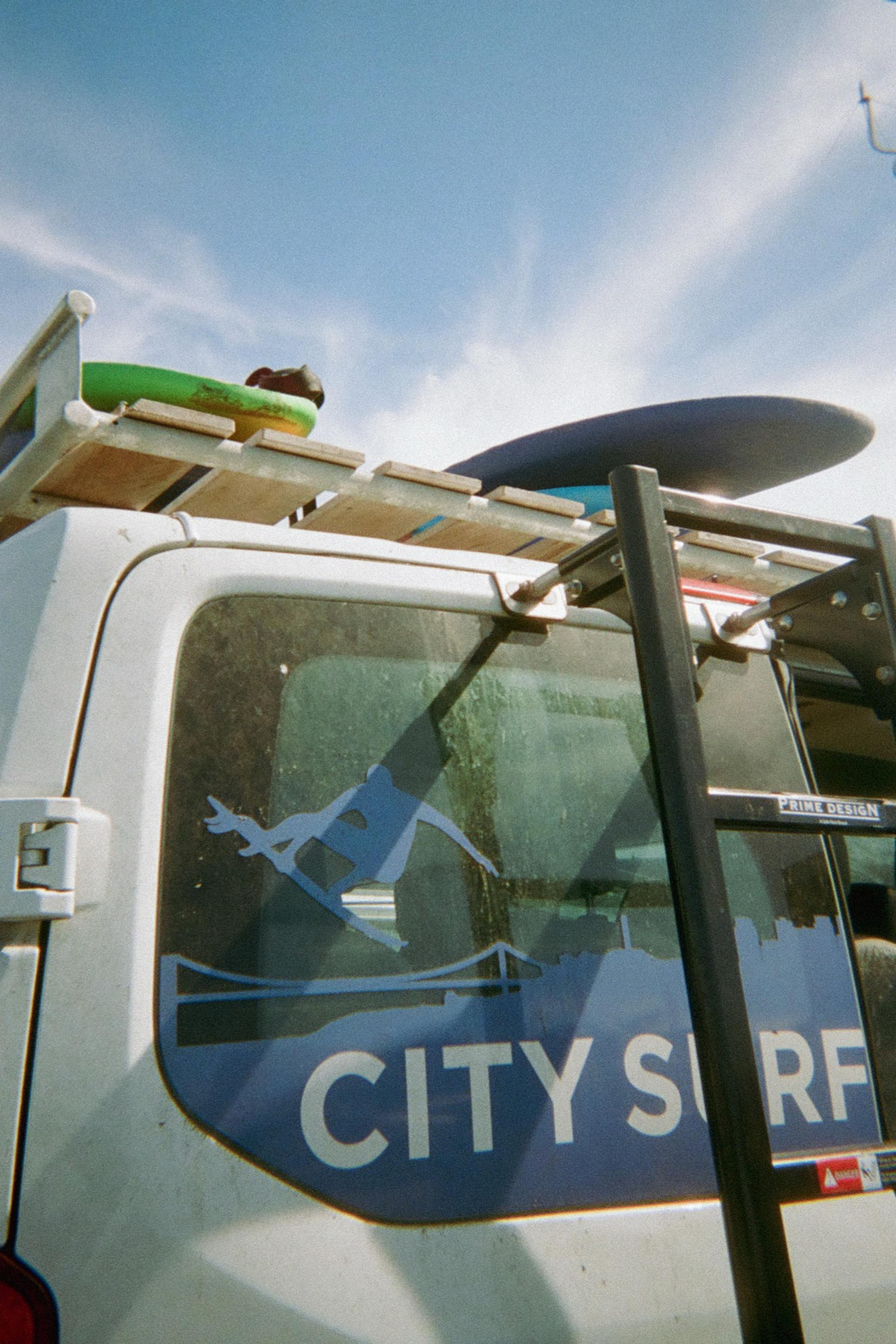 A white van with &quot;City Surf&quot; and a surfing silhouette is shown. Surfboards and a green item are on the roof rack under a clear blue sky.