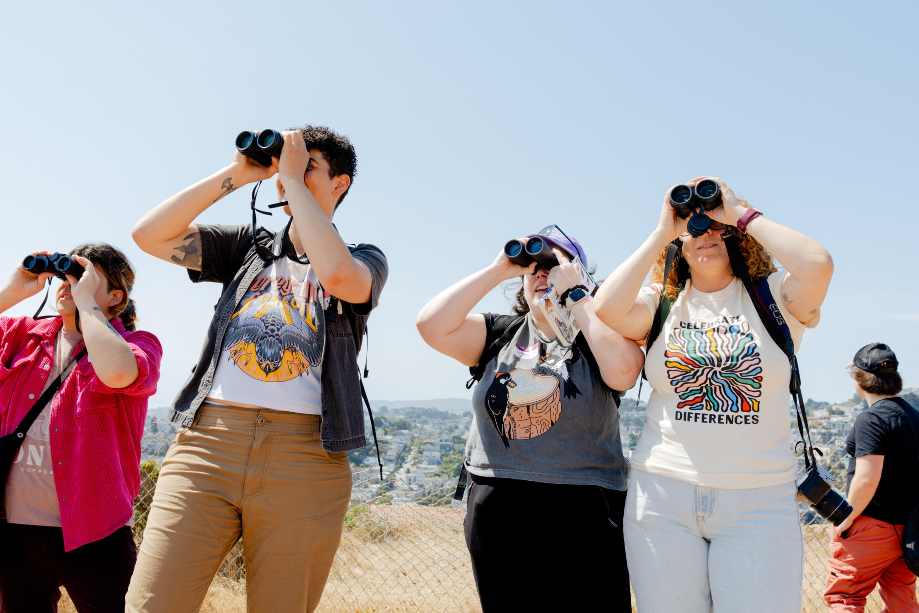 A group of people stands outdoors, looking through binoculars. They are enjoying a sunny day, dressed casually, with city scenery in the background.