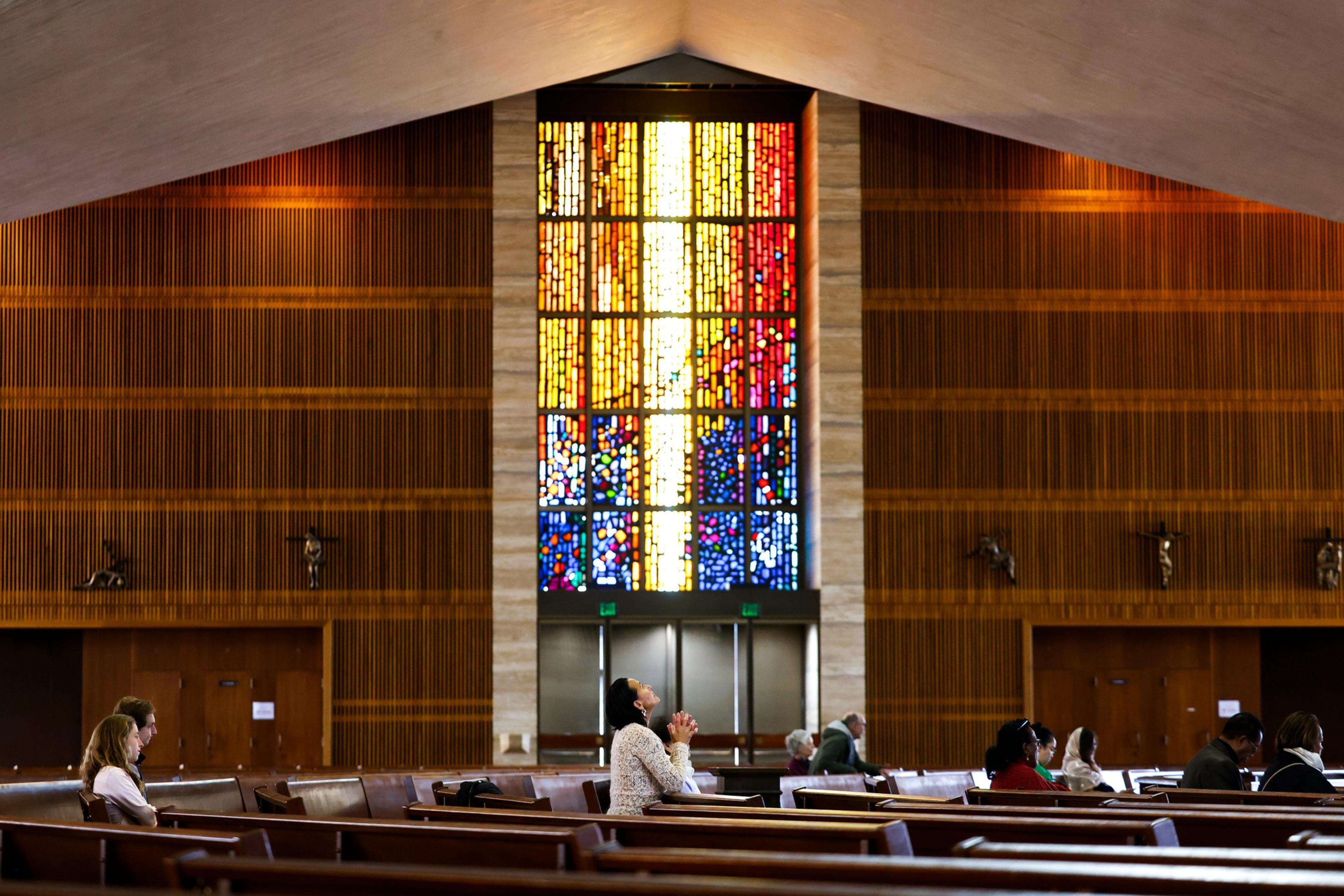 Inside a church, people sit in pews facing a large colorful stained glass window, with one person in white clasping hands as if praying.