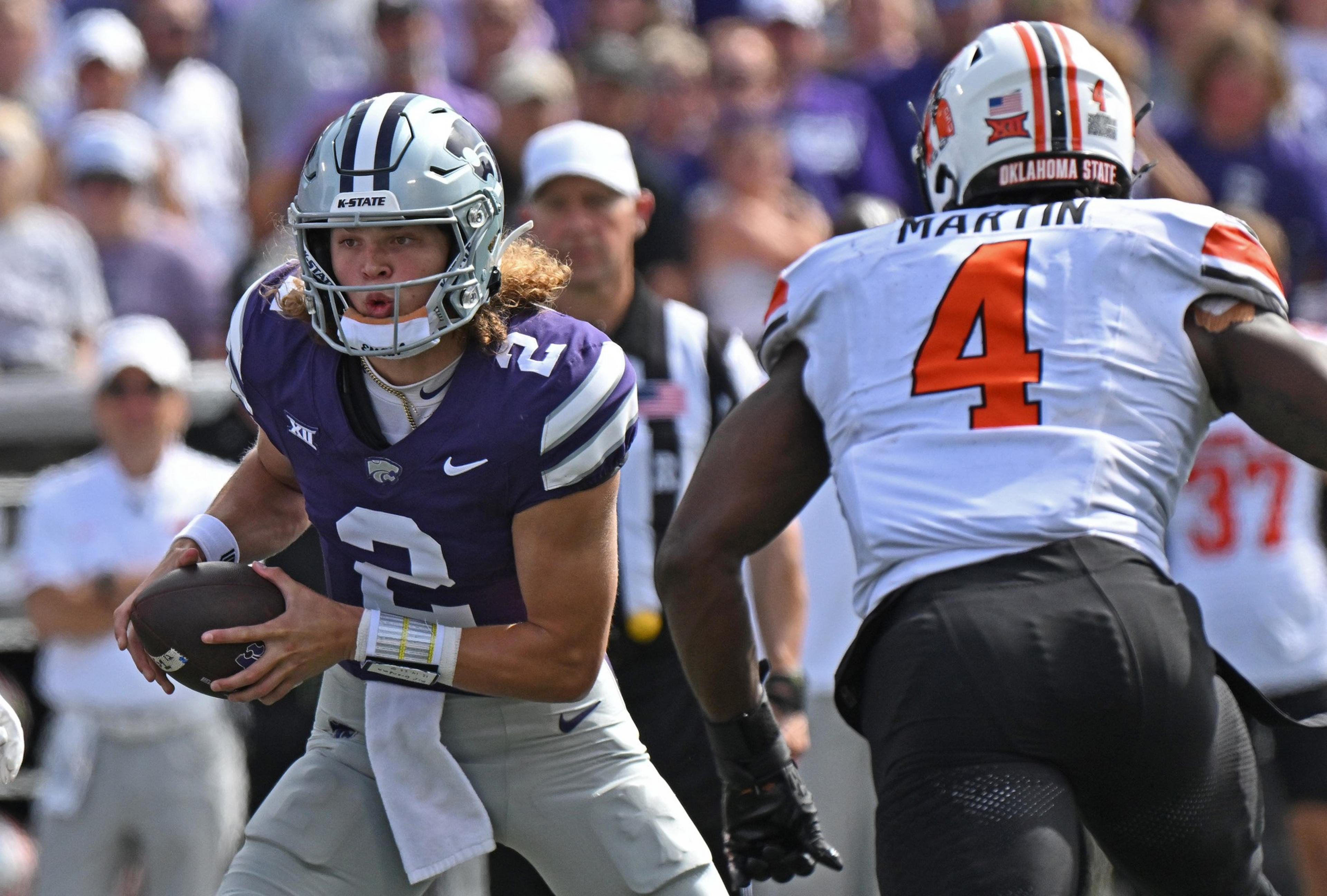A football player in a purple jersey holds the ball, looking to pass, while an opposing player in a white jersey approaches, ready to defend.