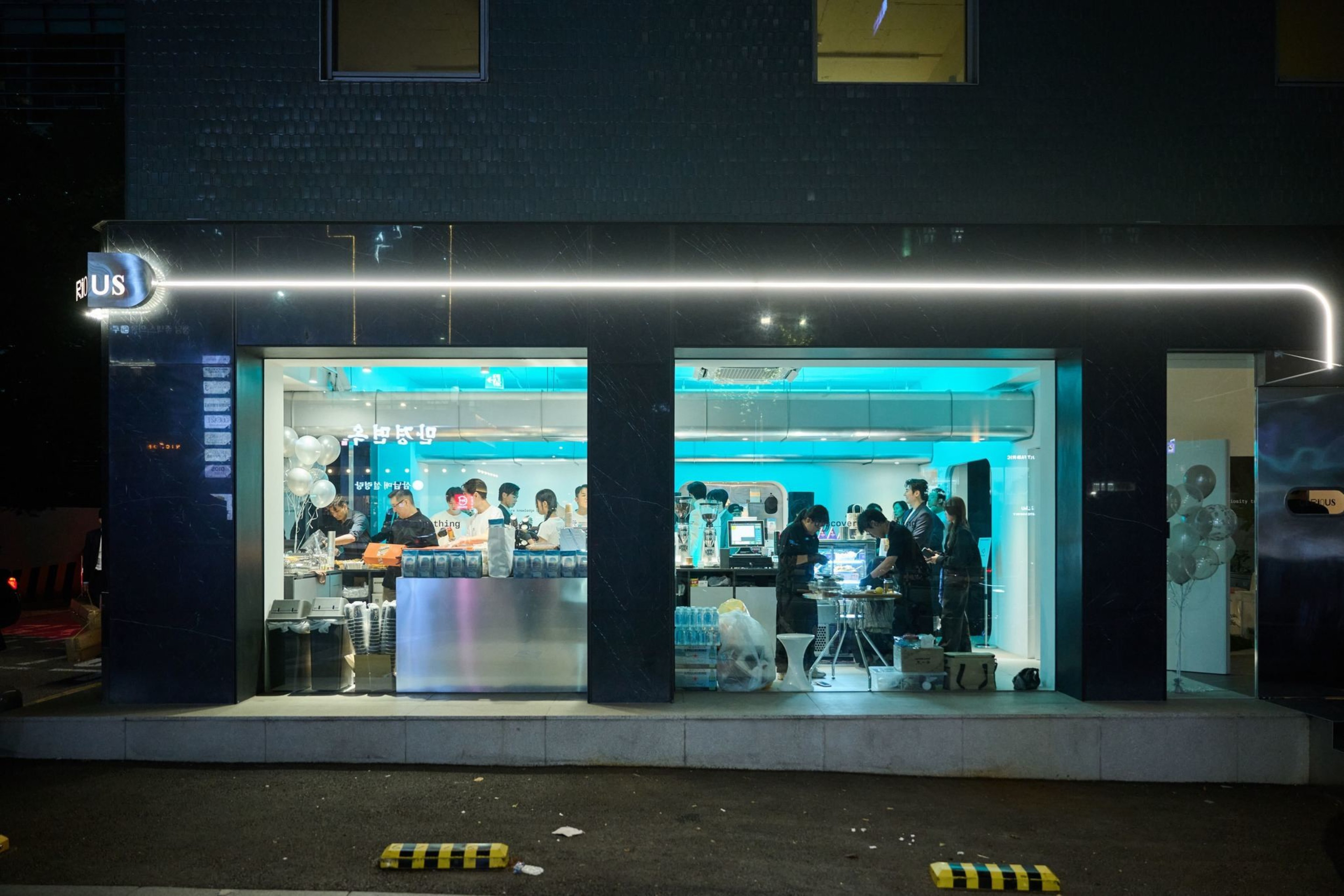 A modern café with bright blue lighting shows several people working behind counters and a group gathered inside, visible through large street-facing windows.