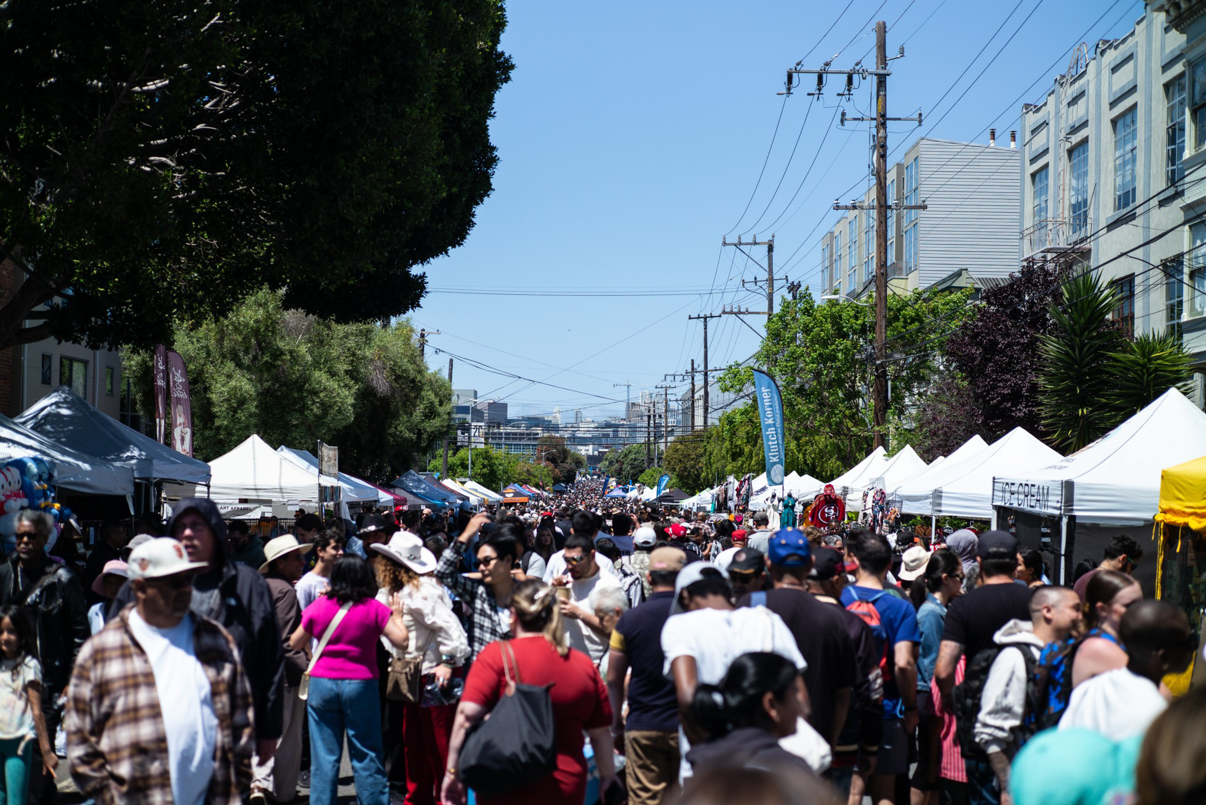 A crowded outdoor street market with people walking between rows of white tents under a clear blue sky and tall buildings on the right.