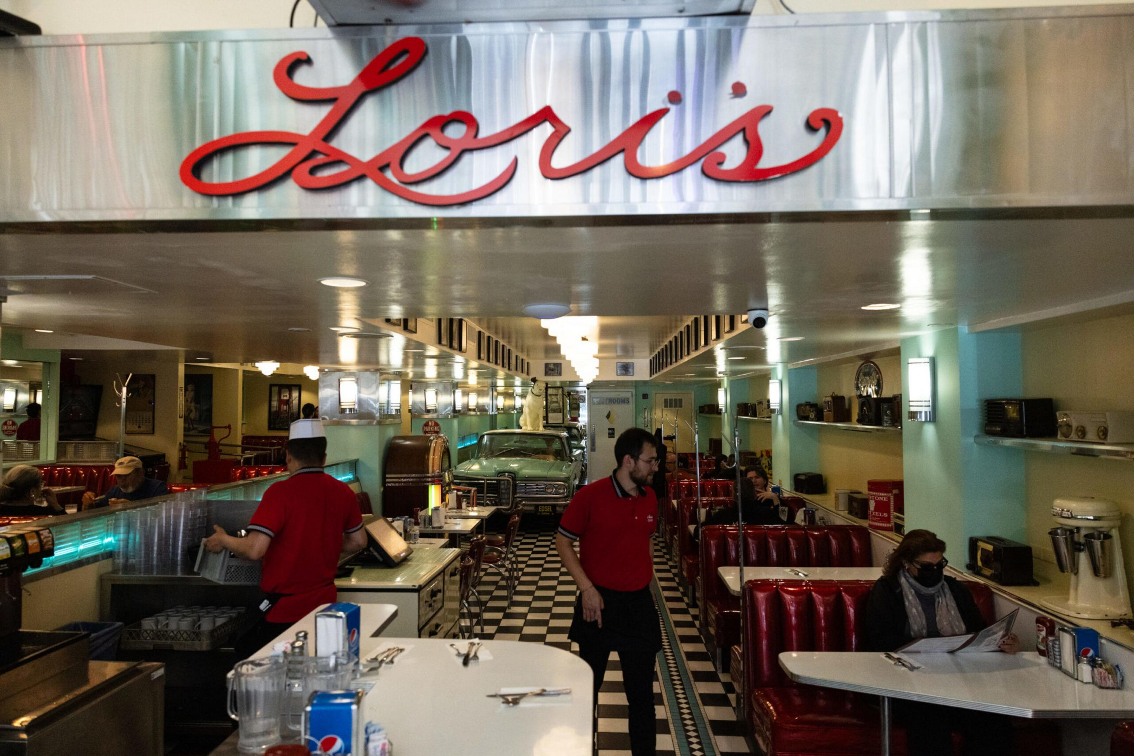 Lori’s diner features red booths, black-and-white checkered floors, a vintage car inside, and staff in red shirts serving customers.