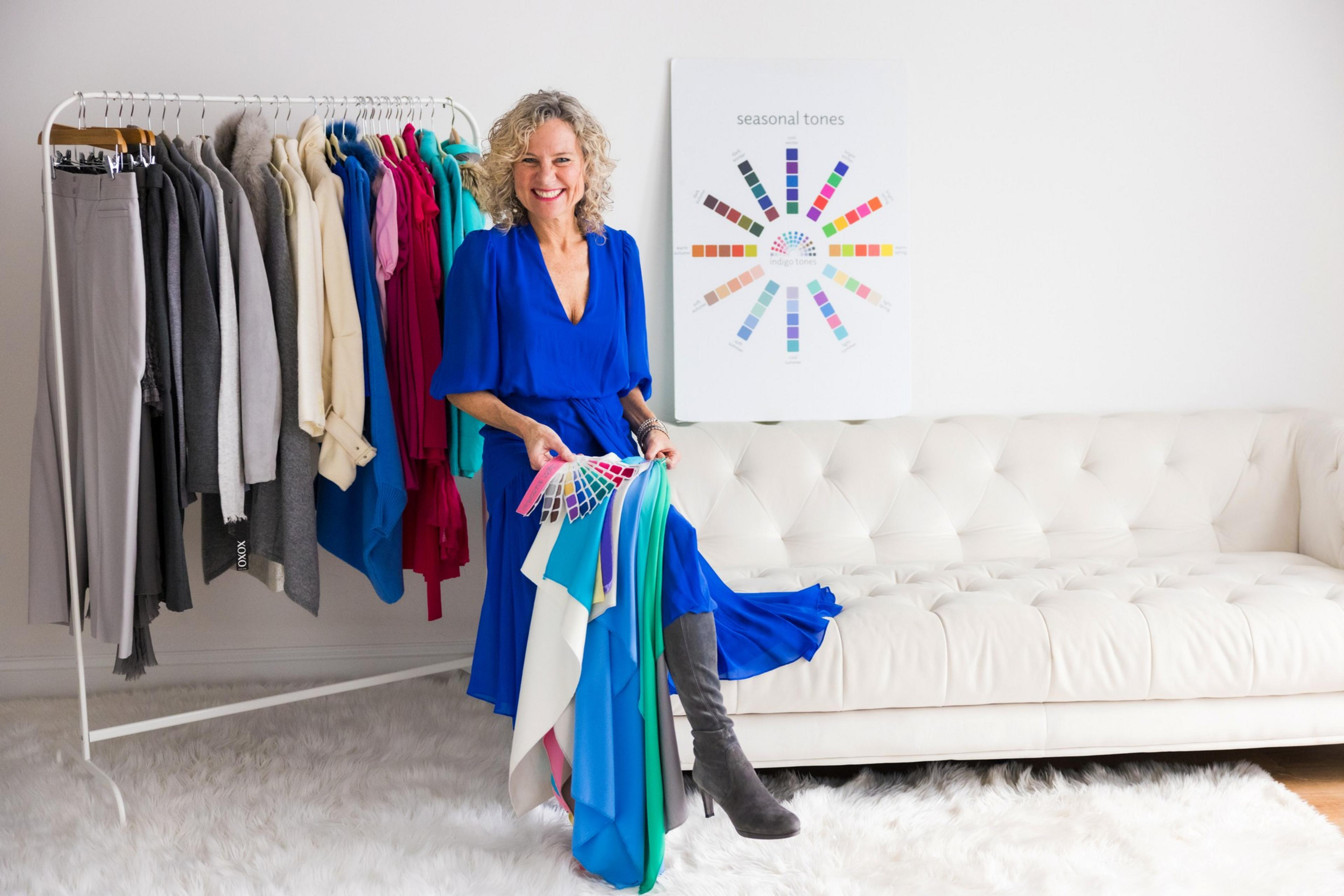 A smiling woman in a blue dress holds fabric samples and stands next to a clothing rack filled with various garments. Behind her is a white sofa and a &quot;seasonal tones&quot; color chart.