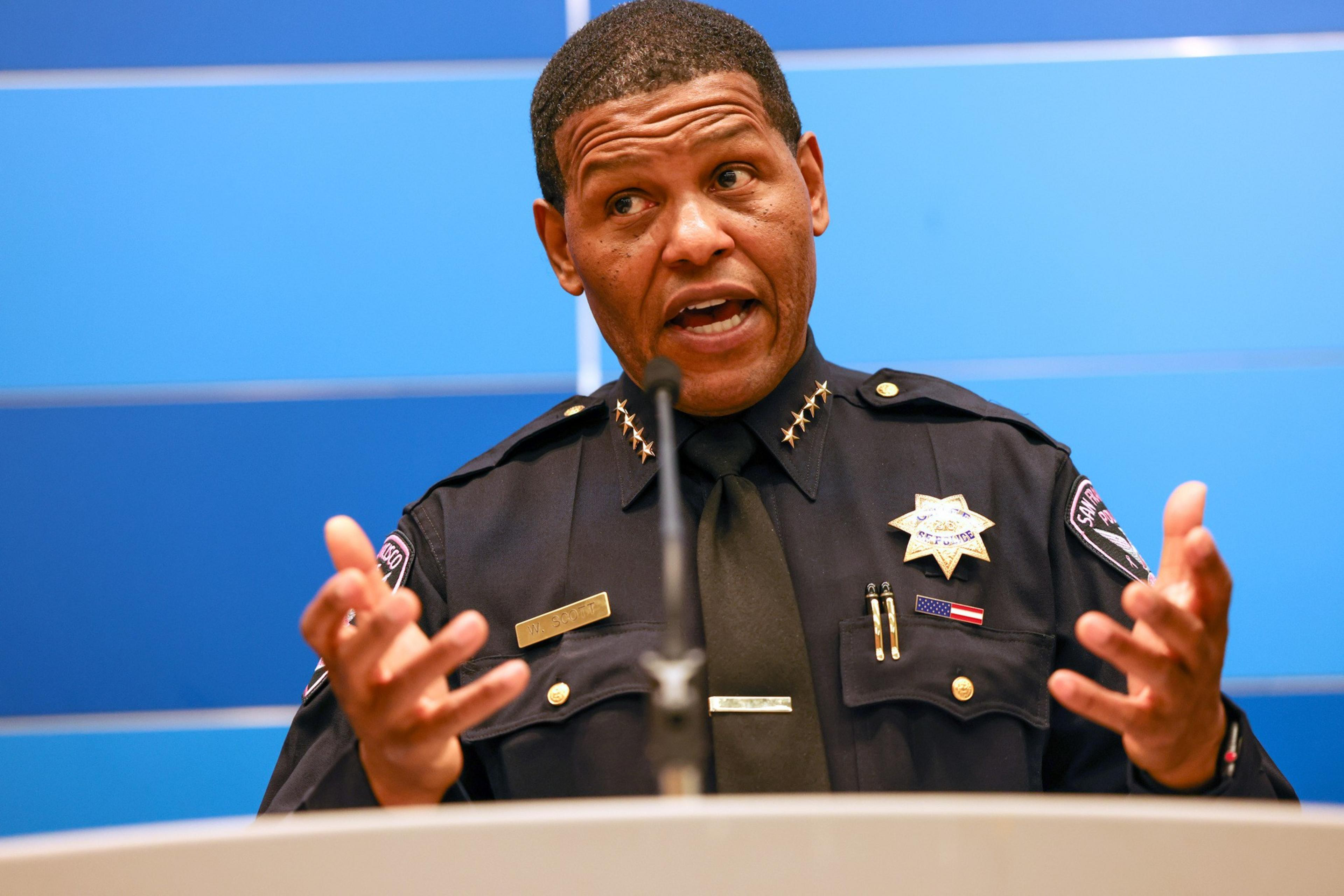 A police officer in uniform speaks passionately at a podium with a microphone, gesturing with both hands against a blue background.