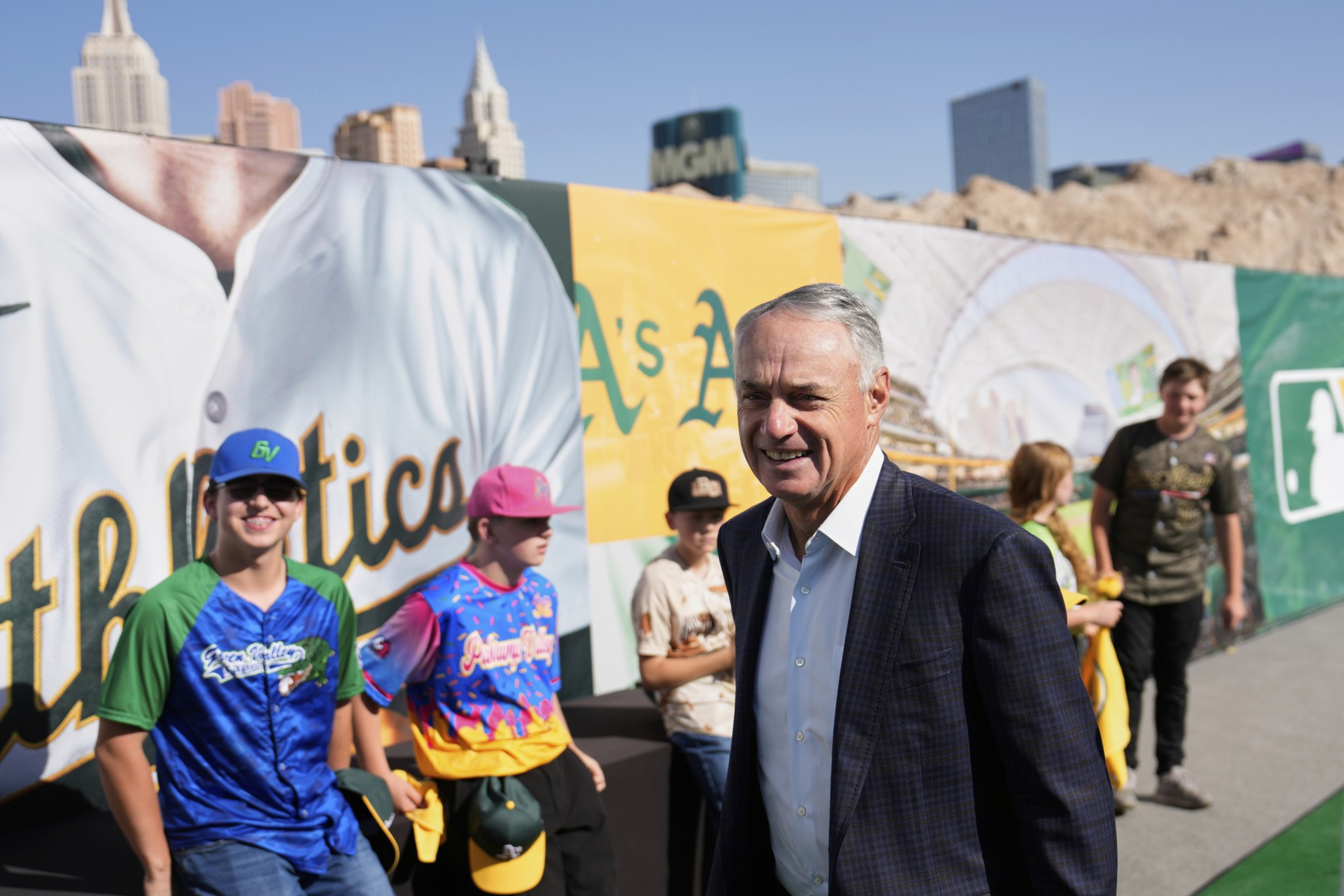 A man in a suit smiles while standing near a colorful baseball-themed backdrop. Behind him, a group of people in baseball jerseys and caps are gathered.