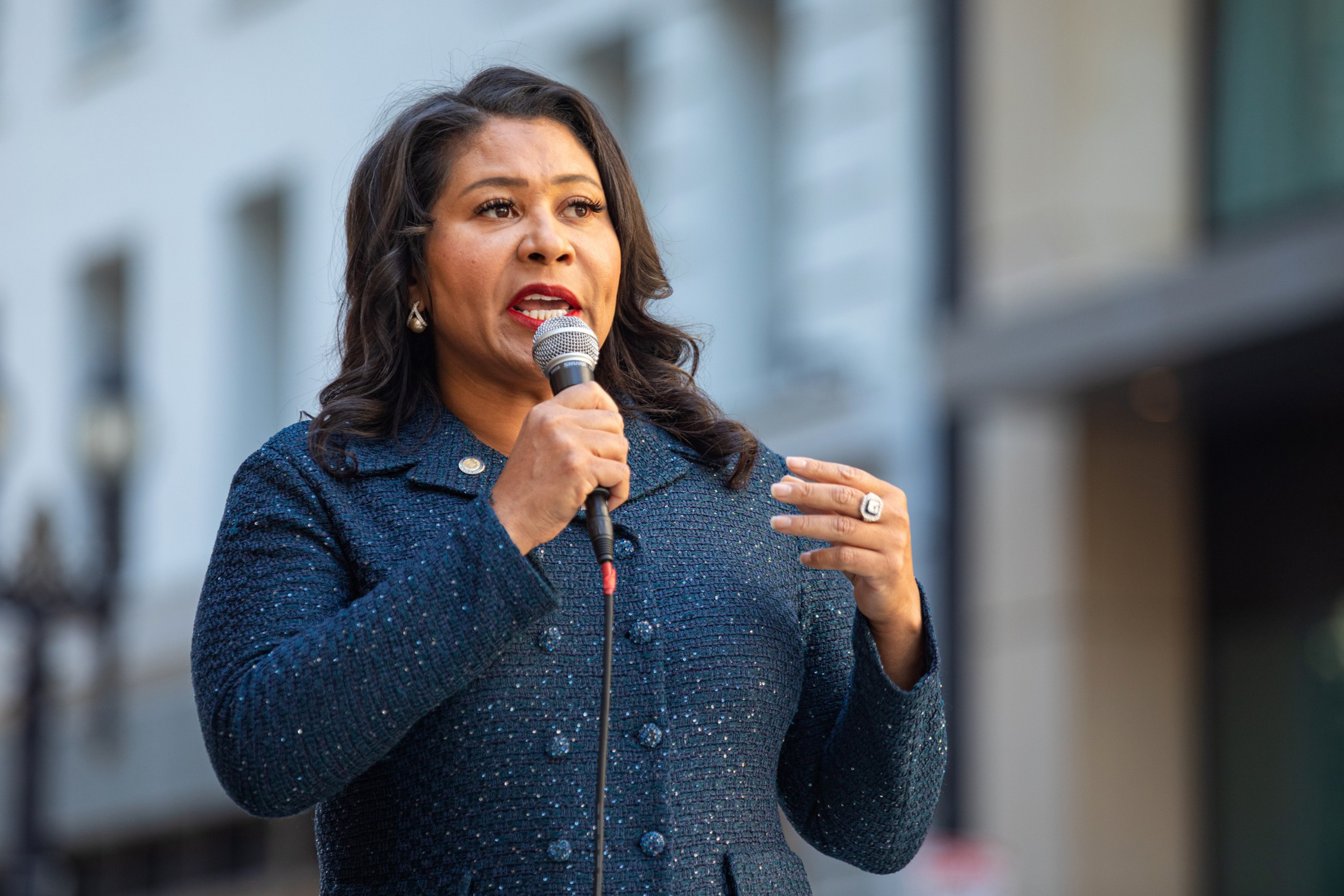 A woman wearing a dark-colored blouse and jacket holds a microphone and addresses an unseen audience.