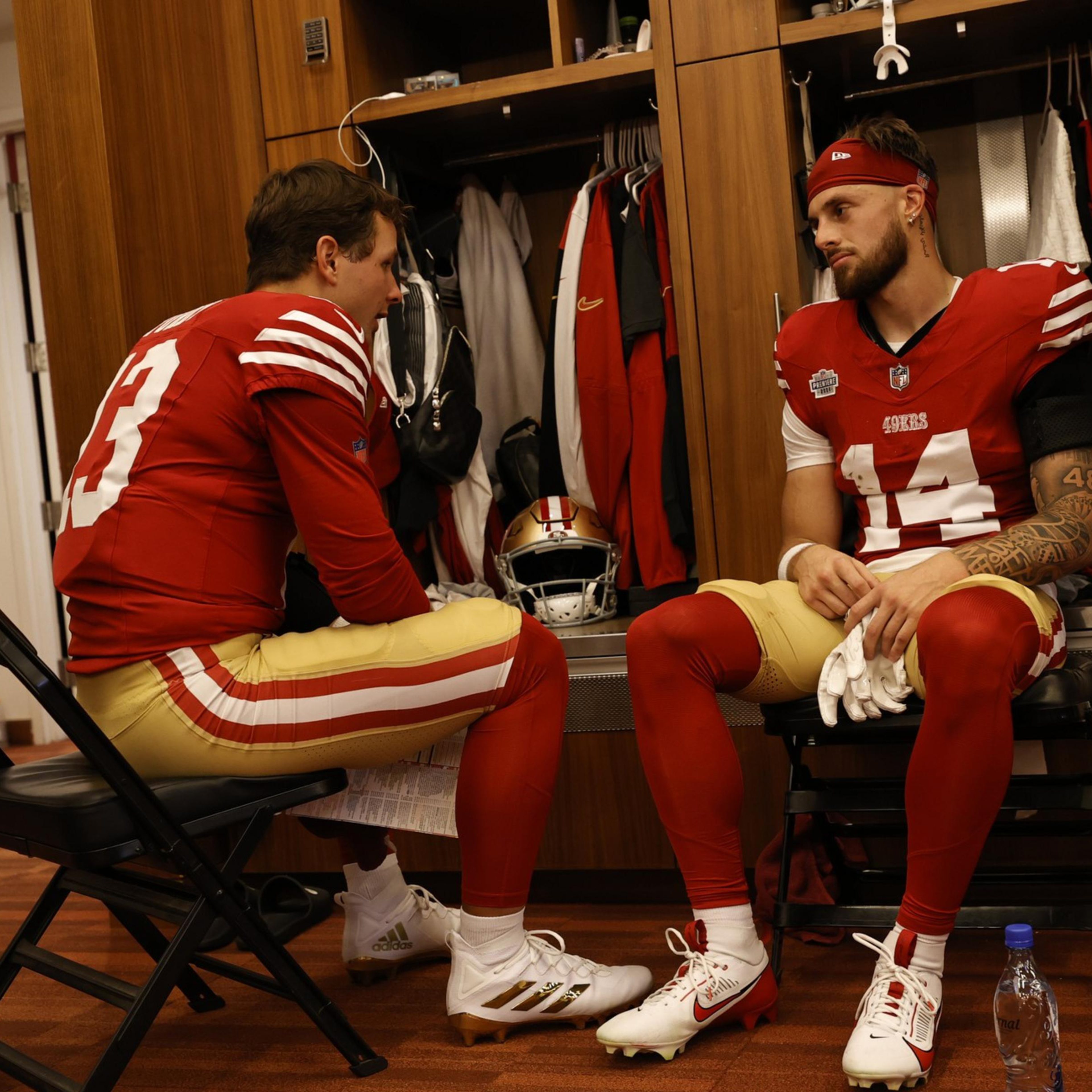 Two football players in red and gold uniforms sit facing each other in a locker room, engaged in a serious conversation.