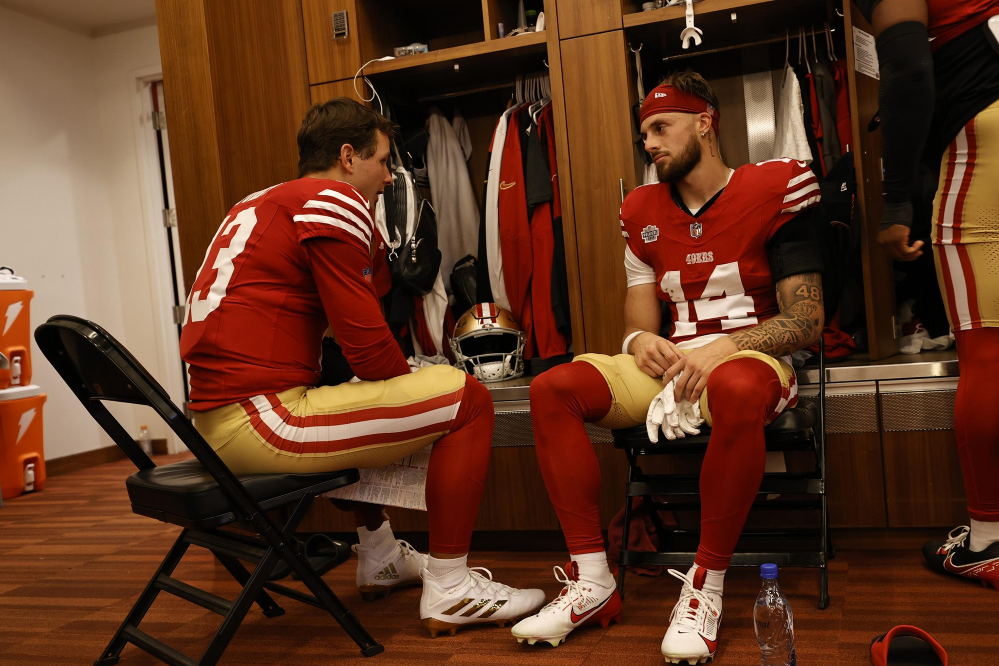 Two football players in red and gold uniforms sit facing each other in a locker room, engaged in a serious conversation.