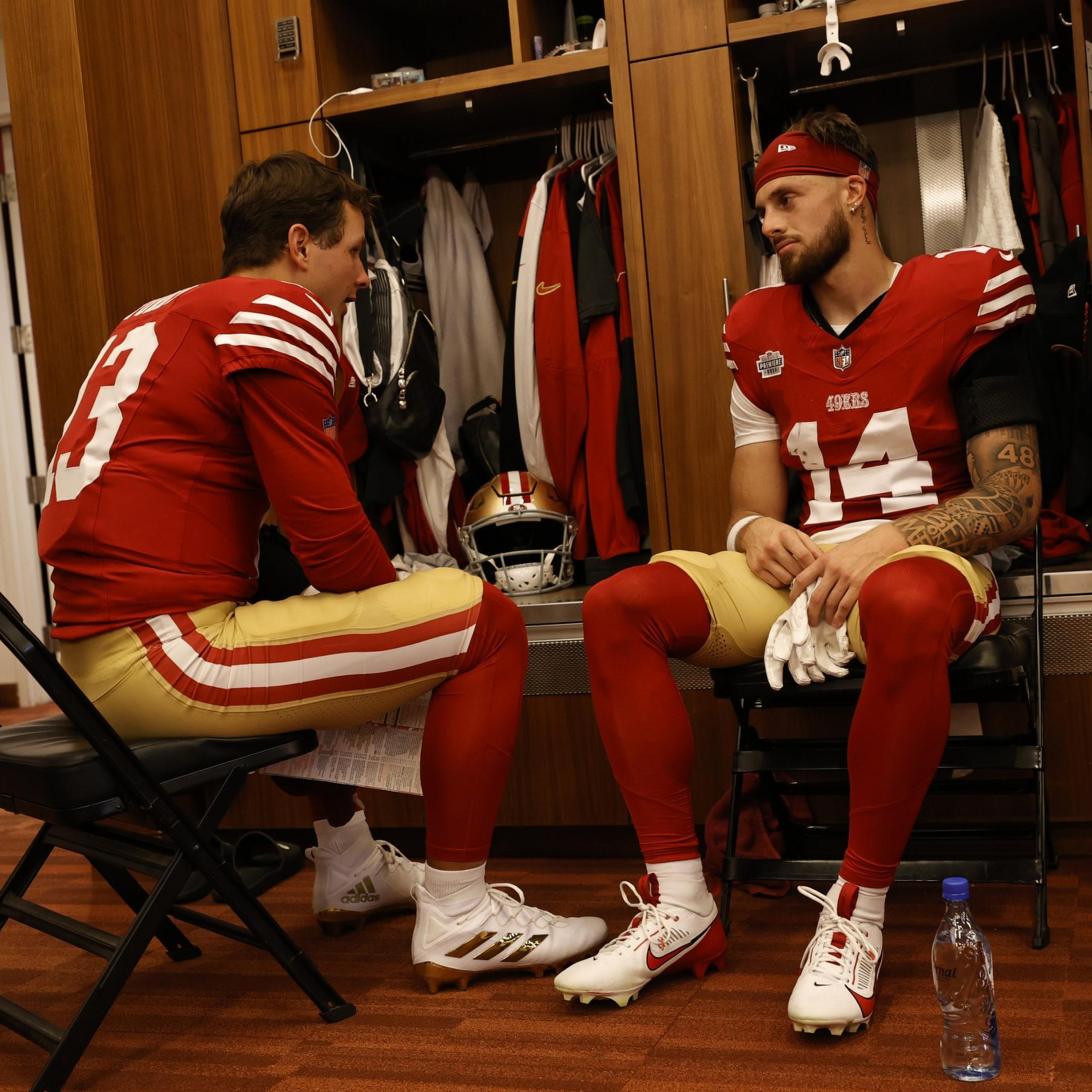 Two football players in red and gold uniforms sit facing each other in a locker room, engaged in a serious conversation.