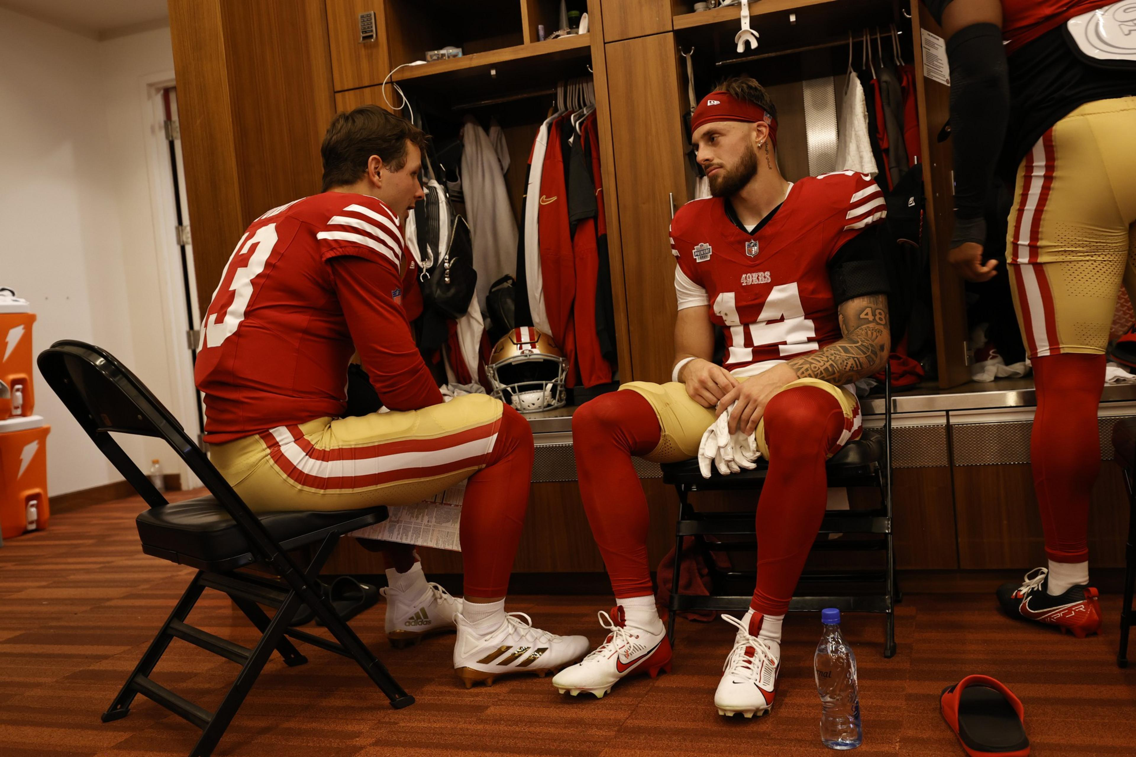 Two football players in red and gold uniforms sit facing each other in a locker room, engaged in a serious conversation.