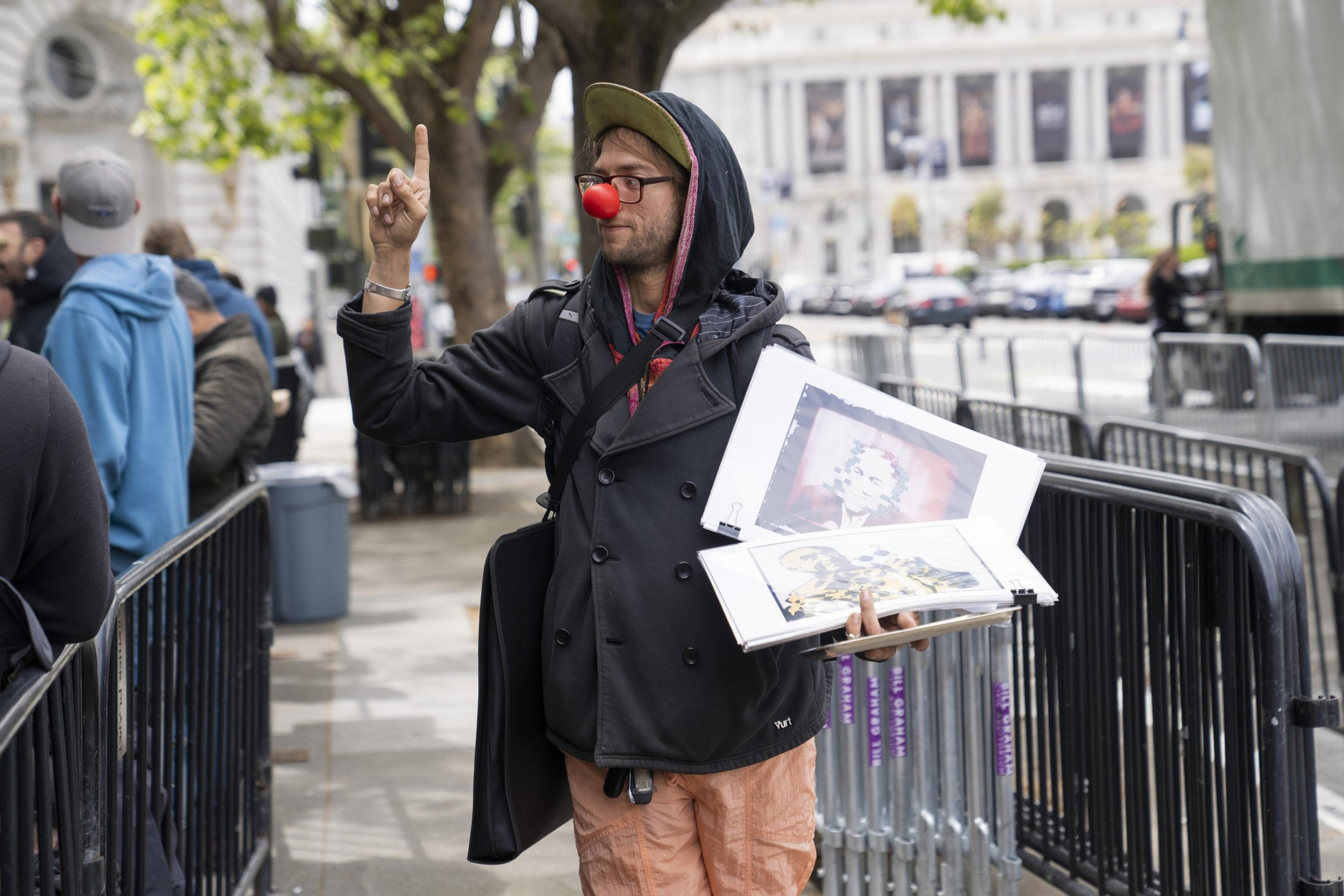 A person wearing a red clown nose and a backpack is holding art prints in one hand and raising the other finger. They're dressed warmly with people around them.