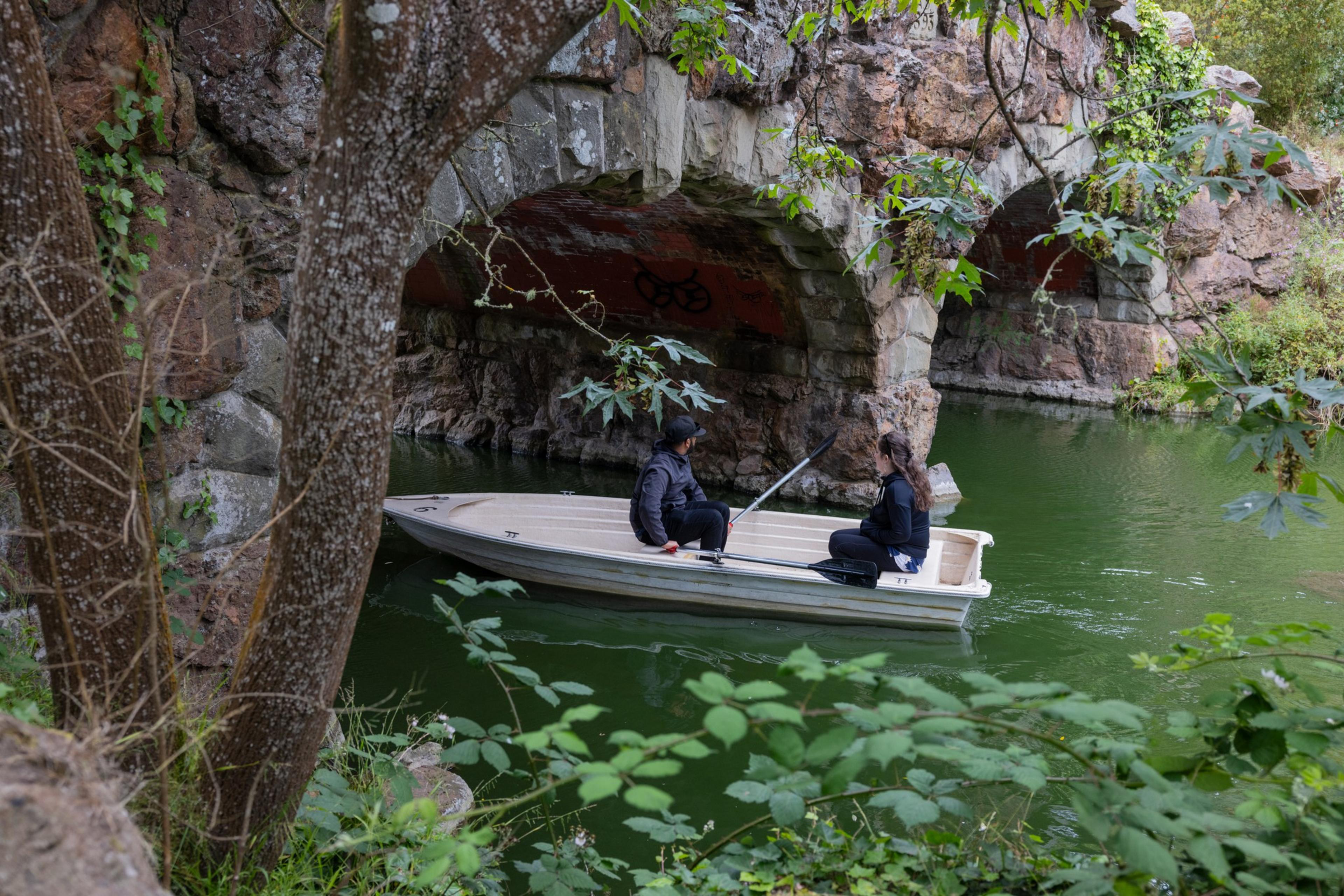 Two people sit in a small white boat rowing under a stone arch bridge surrounded by green water and lush foliage.