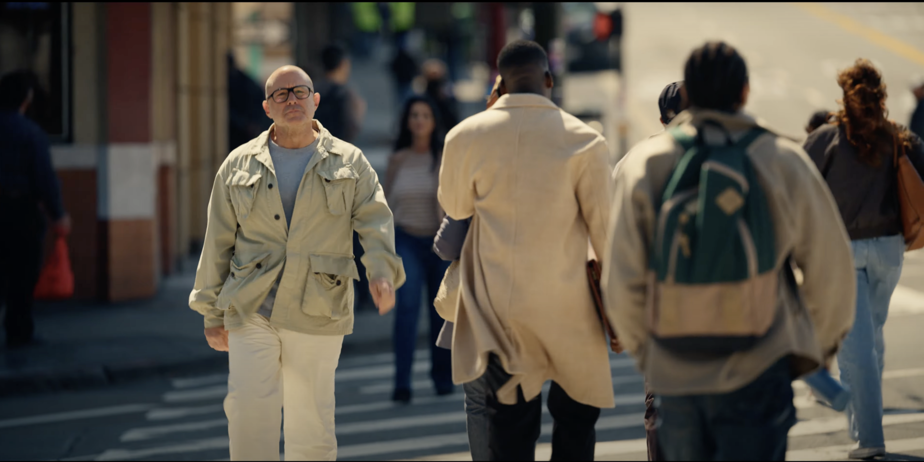 A man in a light jacket walks toward the camera on a busy sidewalk, surrounded by other people going in the opposite direction.