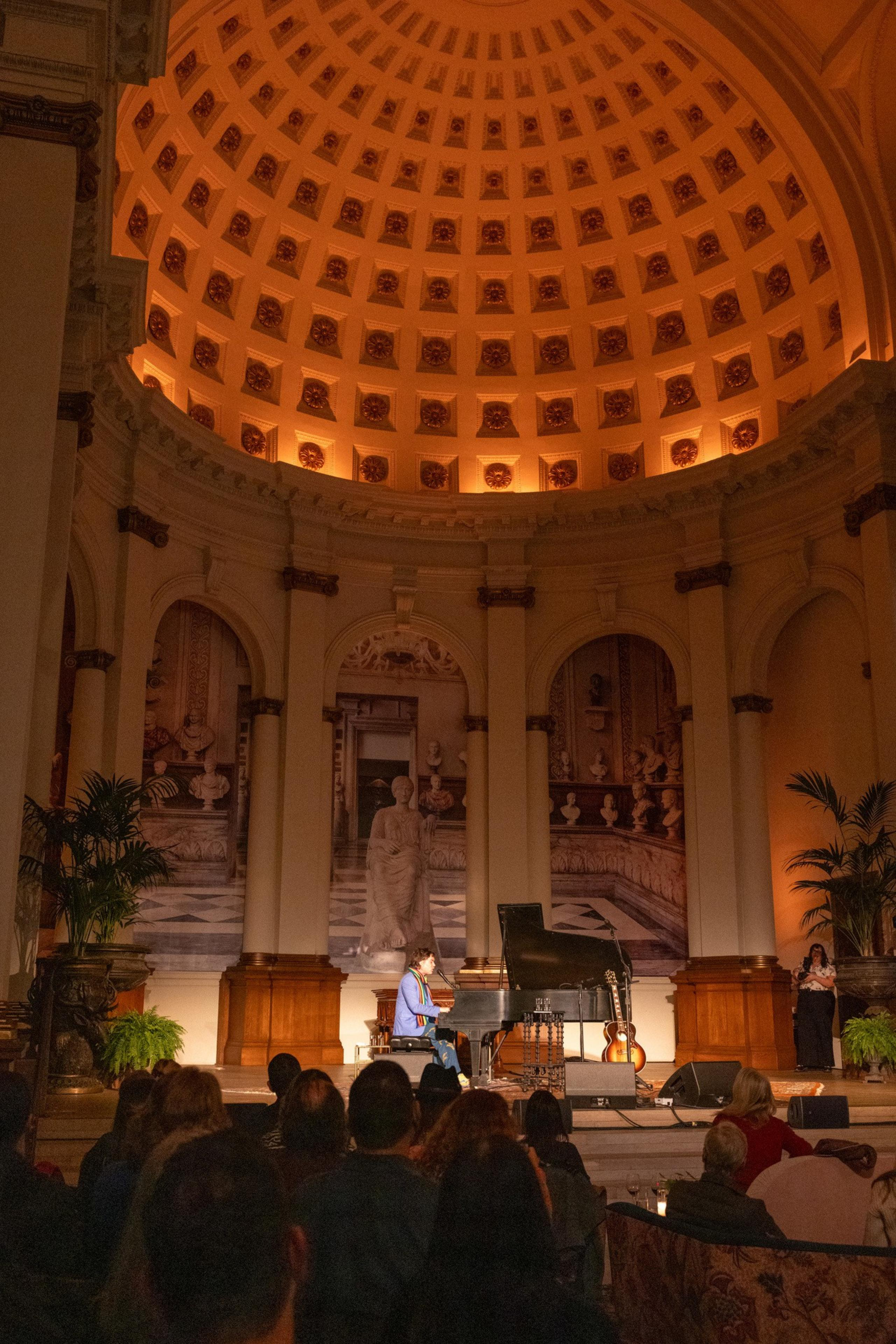 A pianist performs under a grand, ornate dome in a classical hall. The audience watches, surrounded by plants and archways with art.