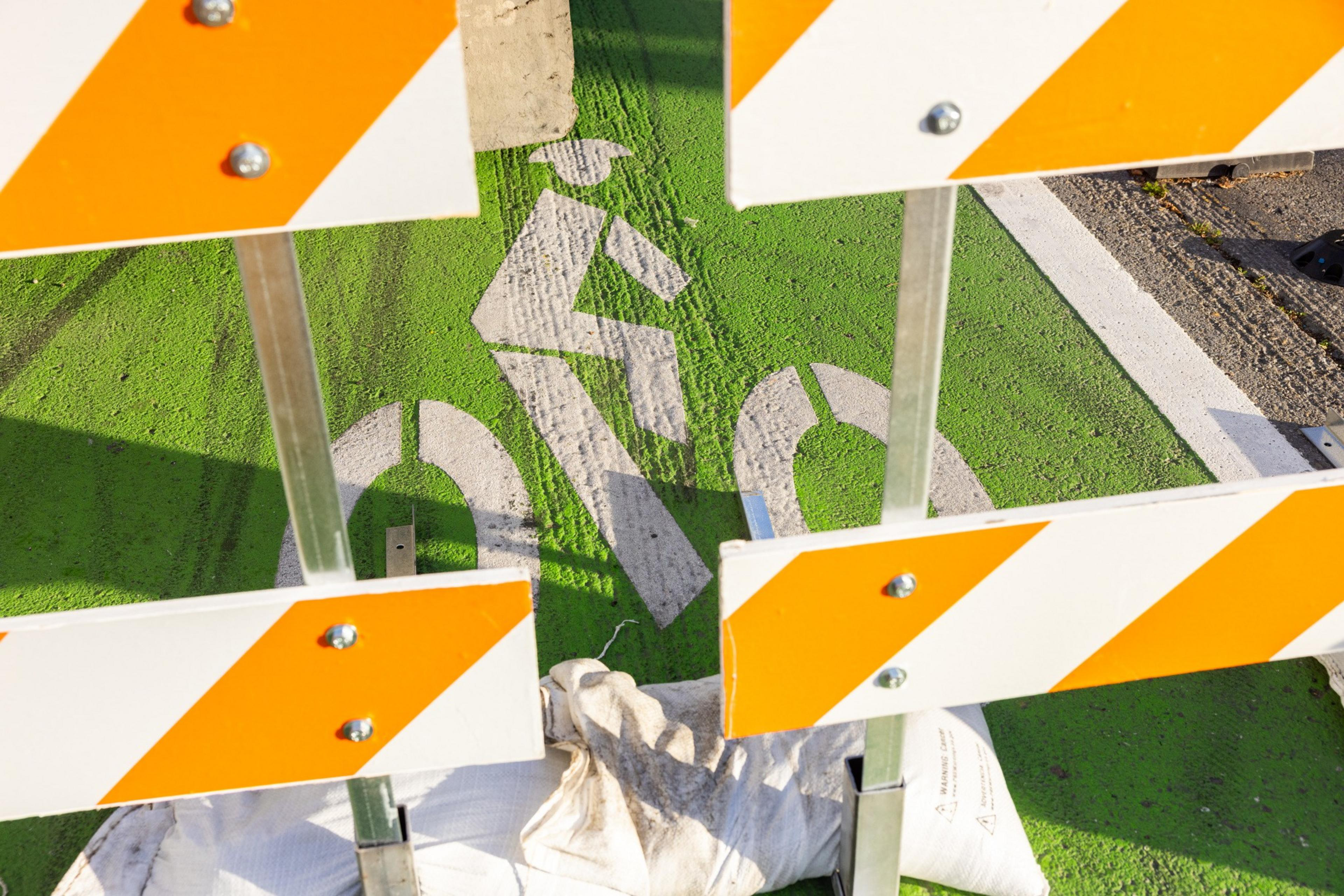 A road barrier with orange and white stripes partially obscures a green bike lane symbol on the ground, indicating a path for cyclists.