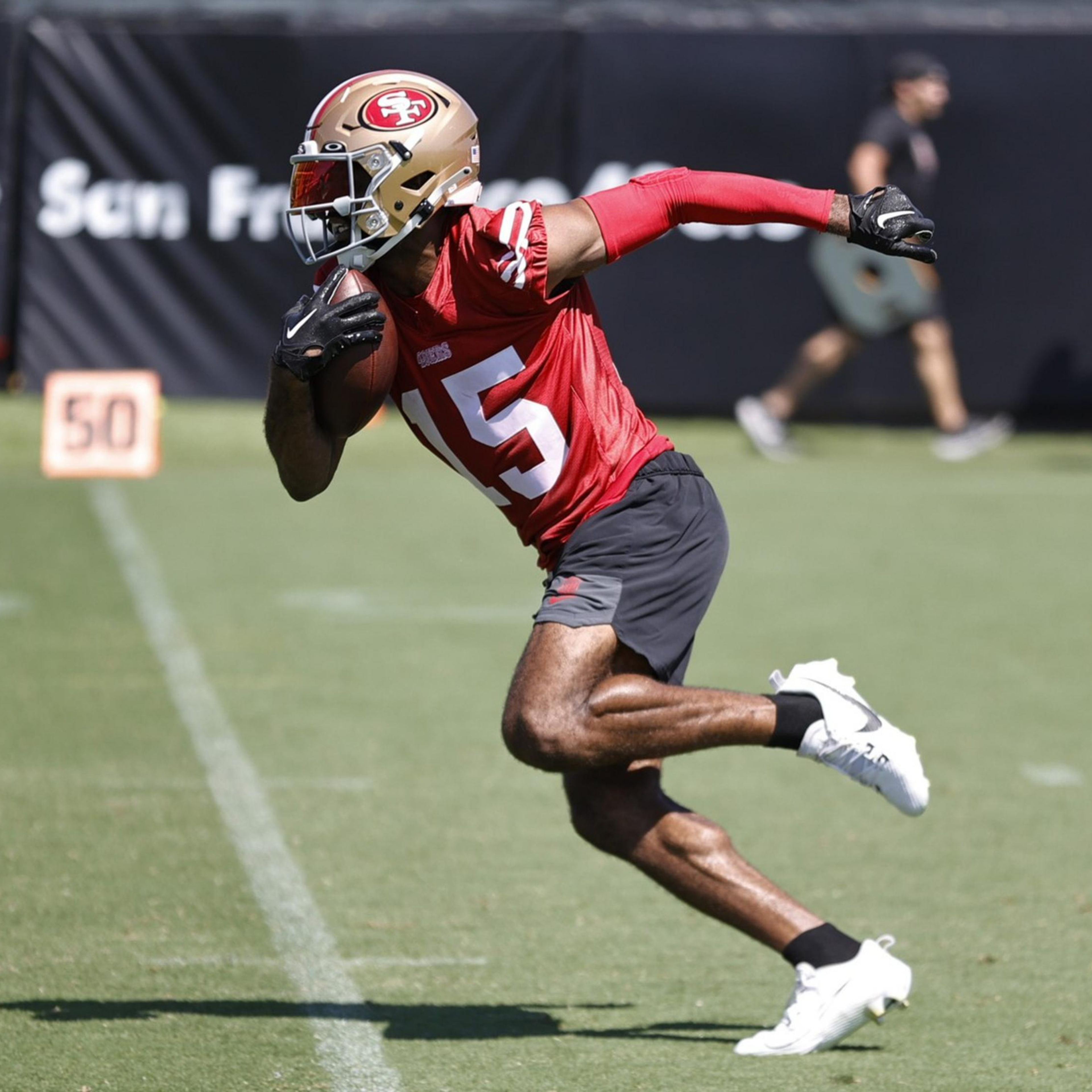 A football player in a red jersey and gold helmet runs on a grassy field holding the ball, with blurred figures and a "50" marker in the background.
