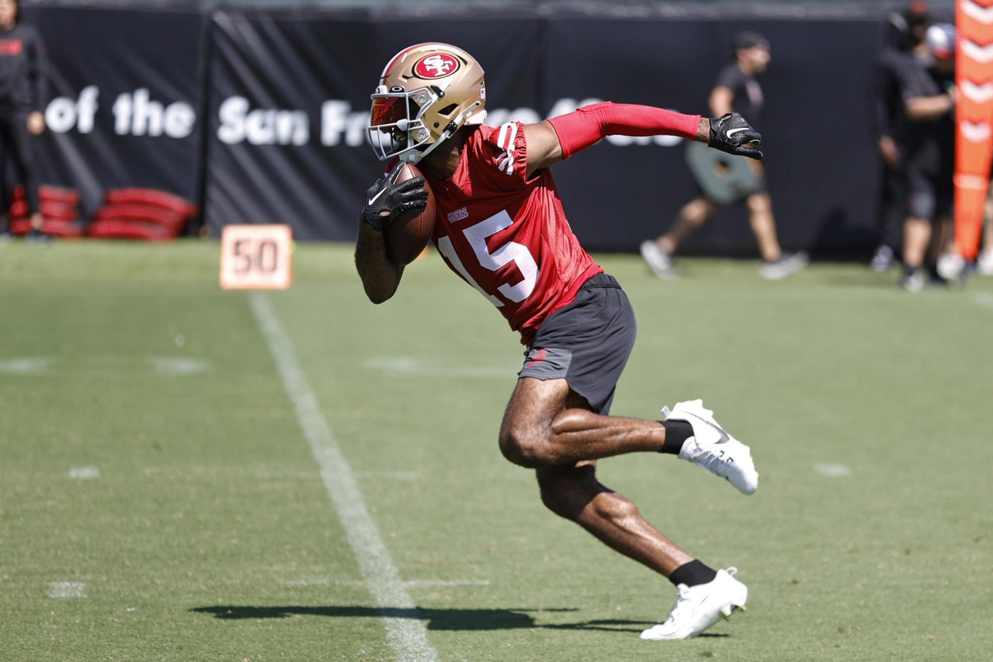 A football player in a red jersey and gold helmet runs on a grassy field holding the ball, with blurred figures and a "50" marker in the background.
