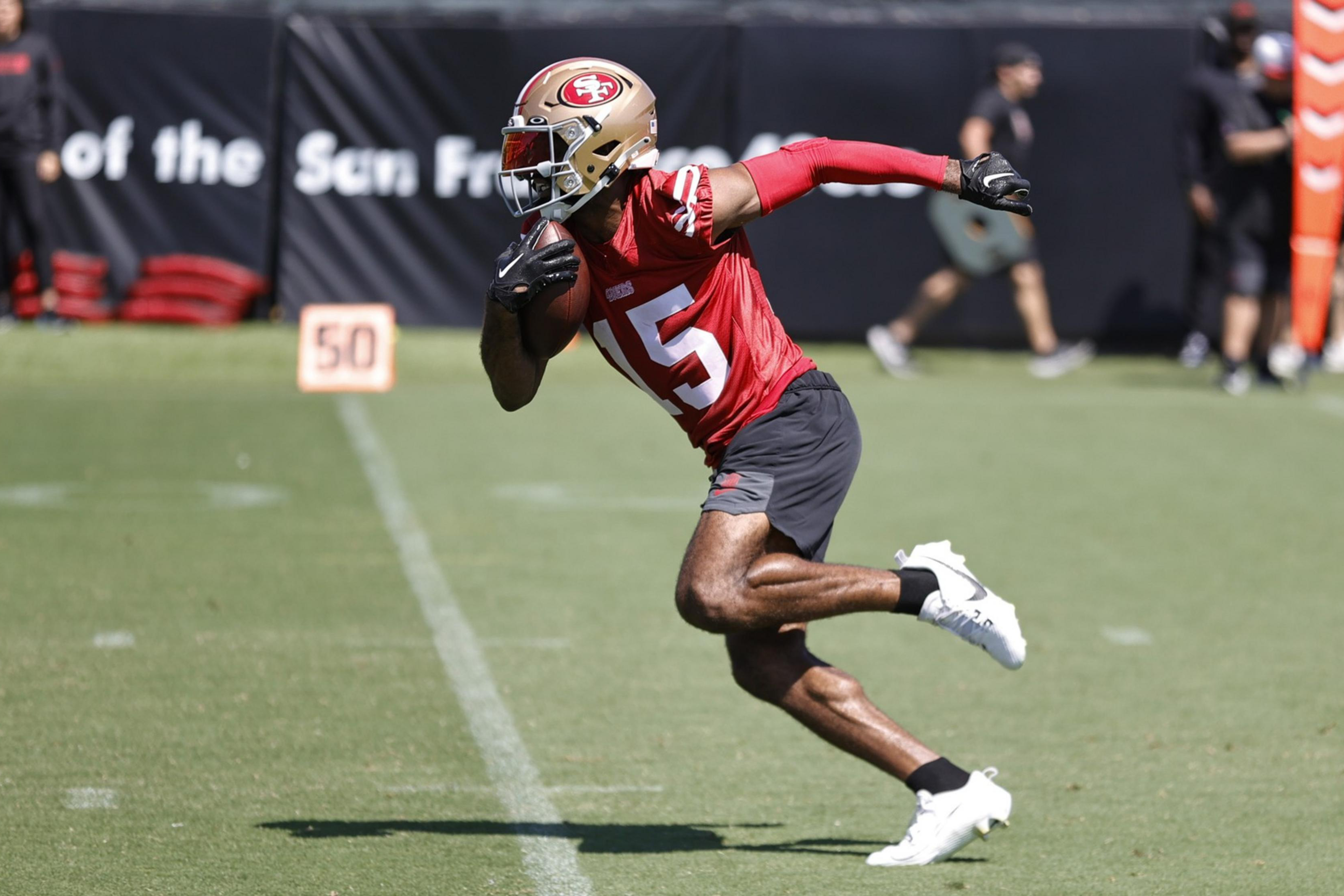 A football player in a red jersey and gold helmet runs on a grassy field holding the ball, with blurred figures and a "50" marker in the background.