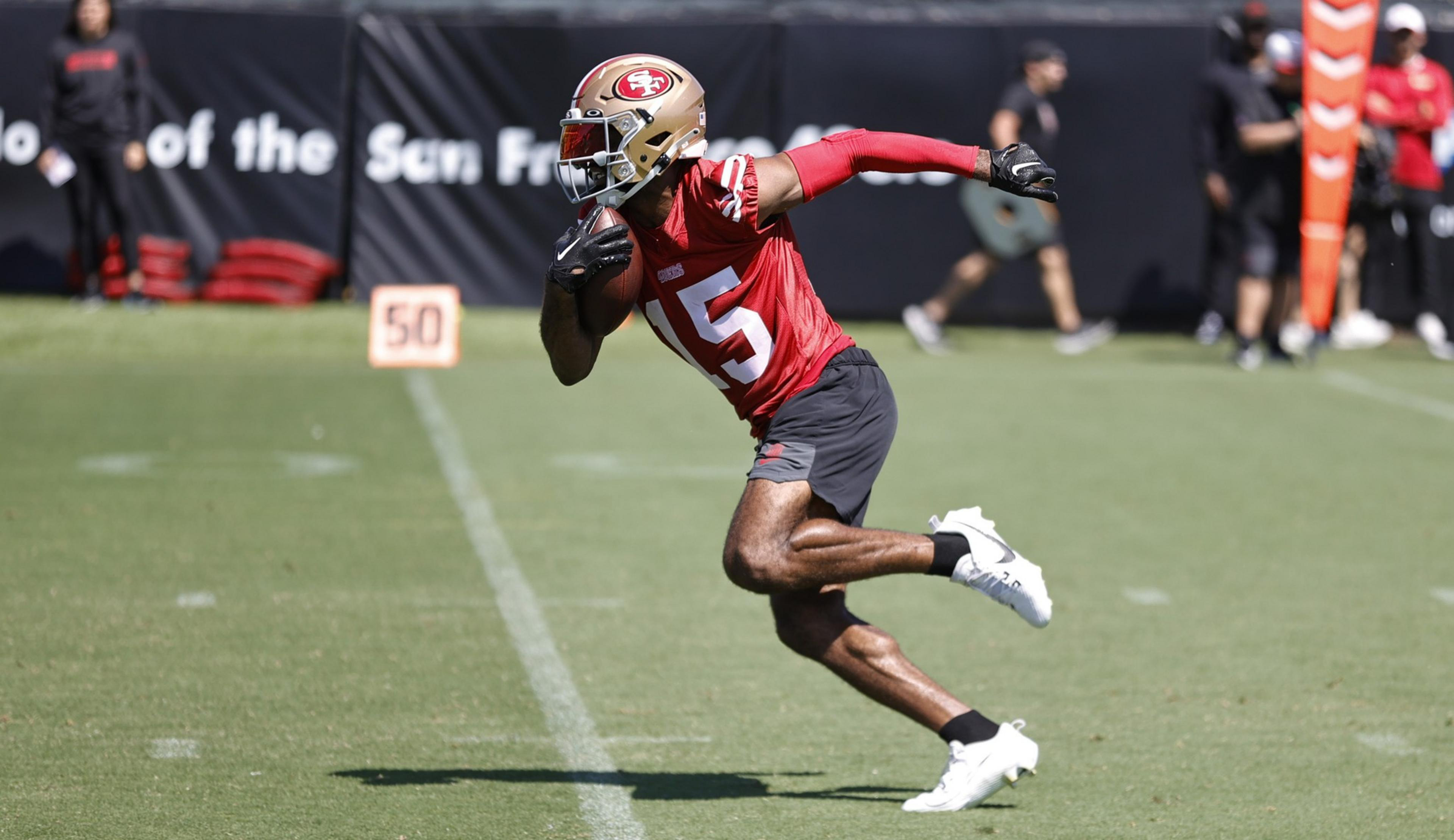A football player in a red jersey and gold helmet runs on a grassy field holding the ball, with blurred figures and a "50" marker in the background.