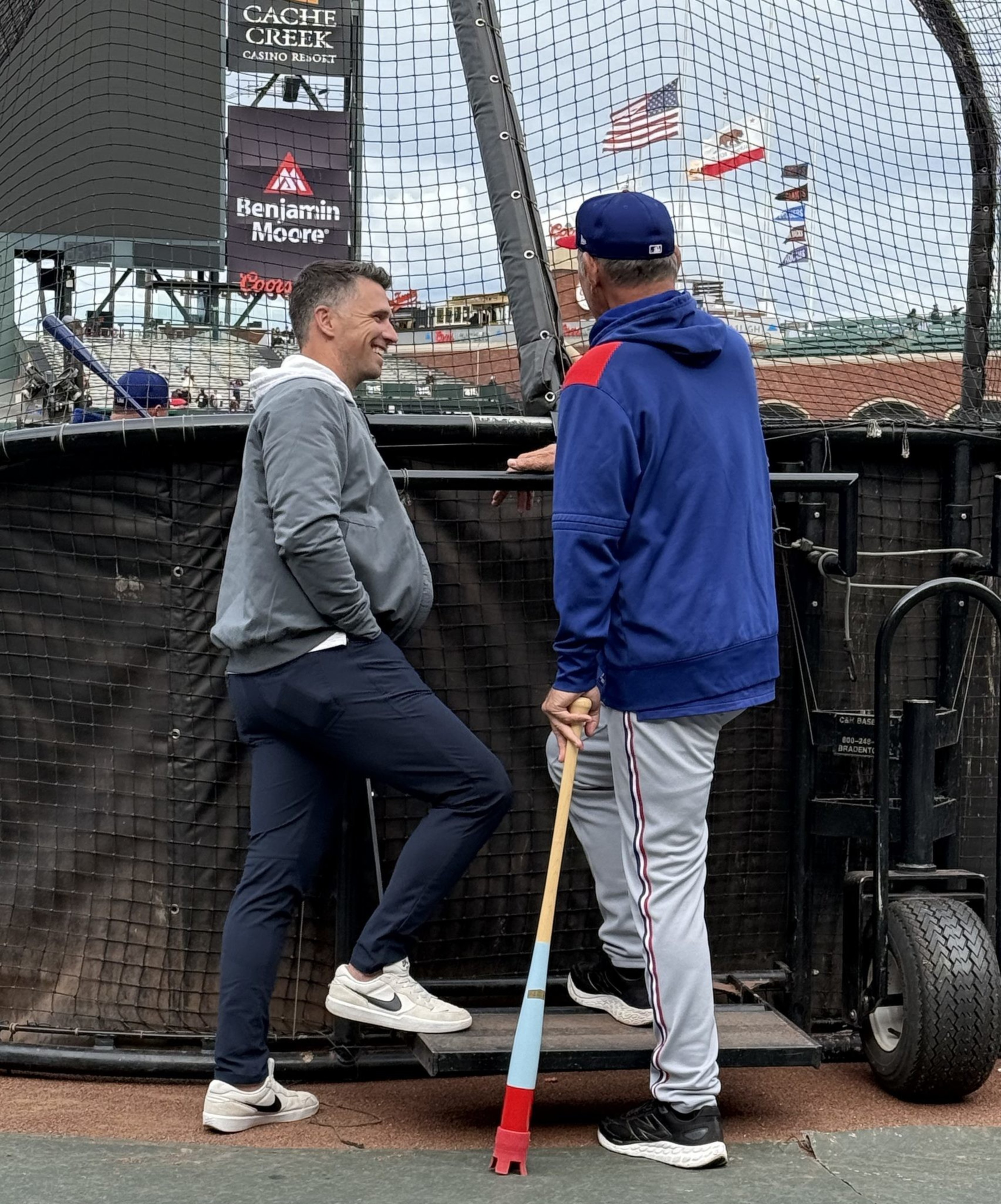 Two men stand near a baseball field's batting cage, chatting. One wears casual attire, the other is in a baseball uniform holding a red and blue bat.