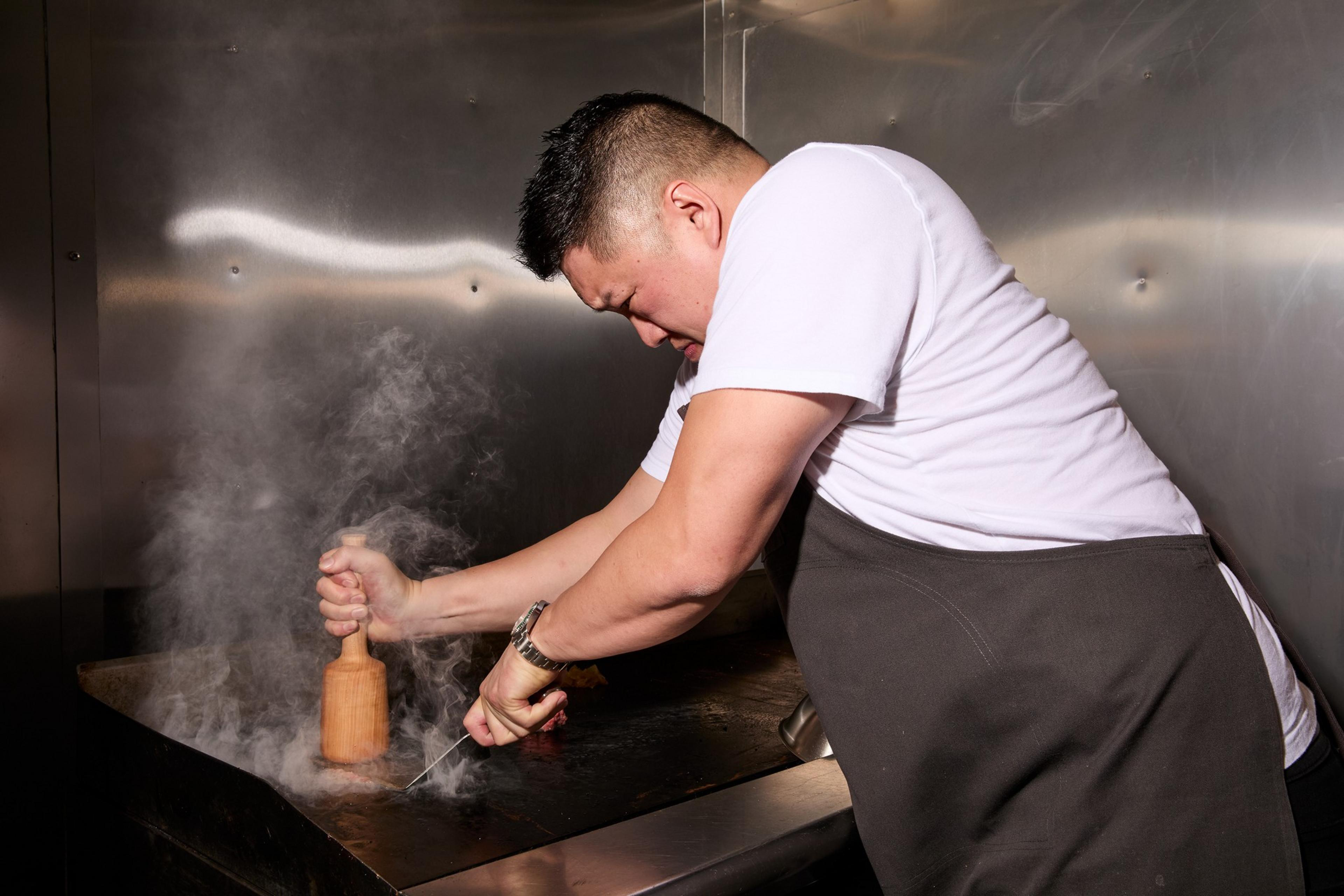 A person in a white shirt and apron presses a wooden mallet onto a hot, smoky grill, with a focused expression.