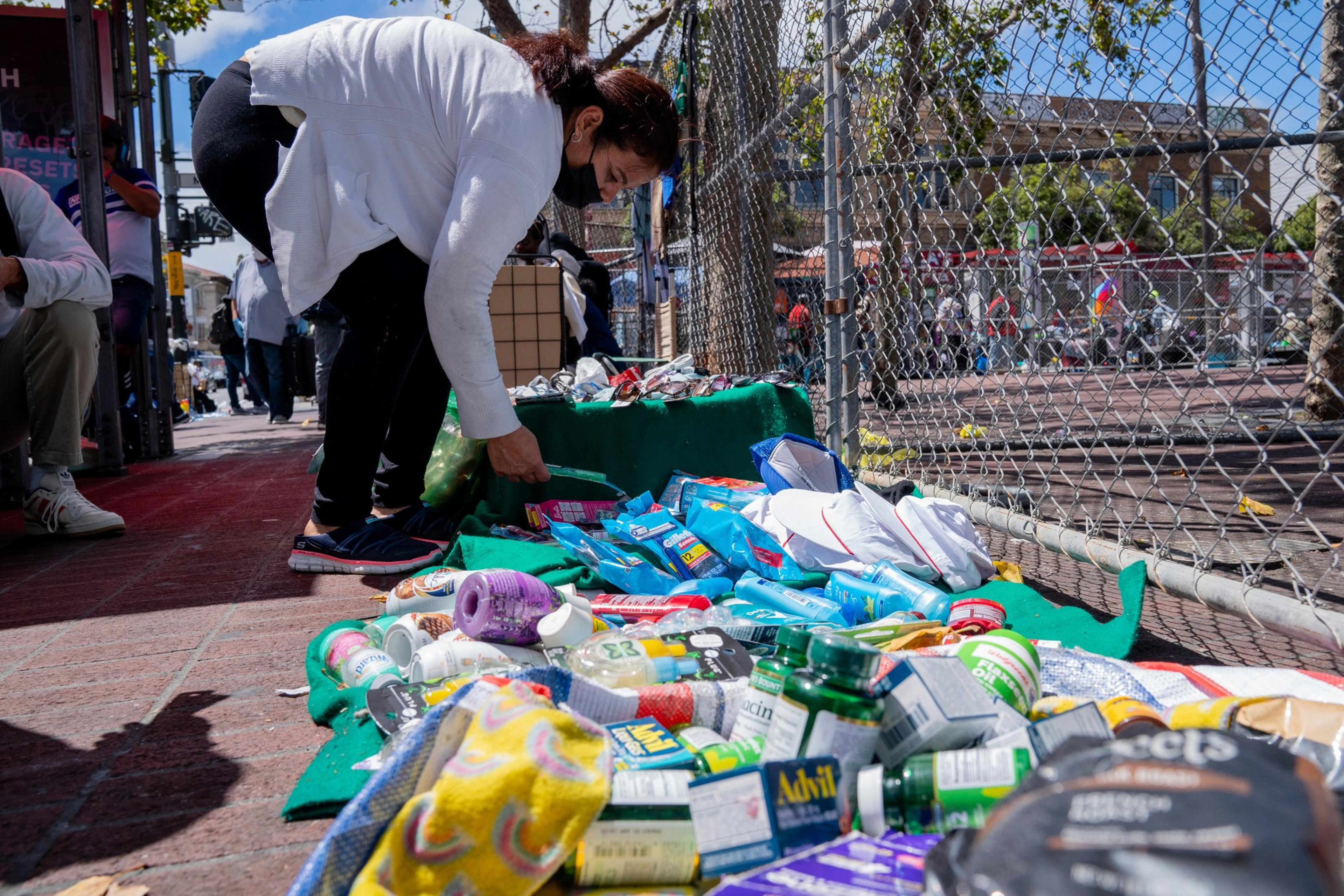 A person in a white jacket and black mask is bending over a sidewalk display of assorted medical supplies and toiletries near a fenced area, with a few people in the background.