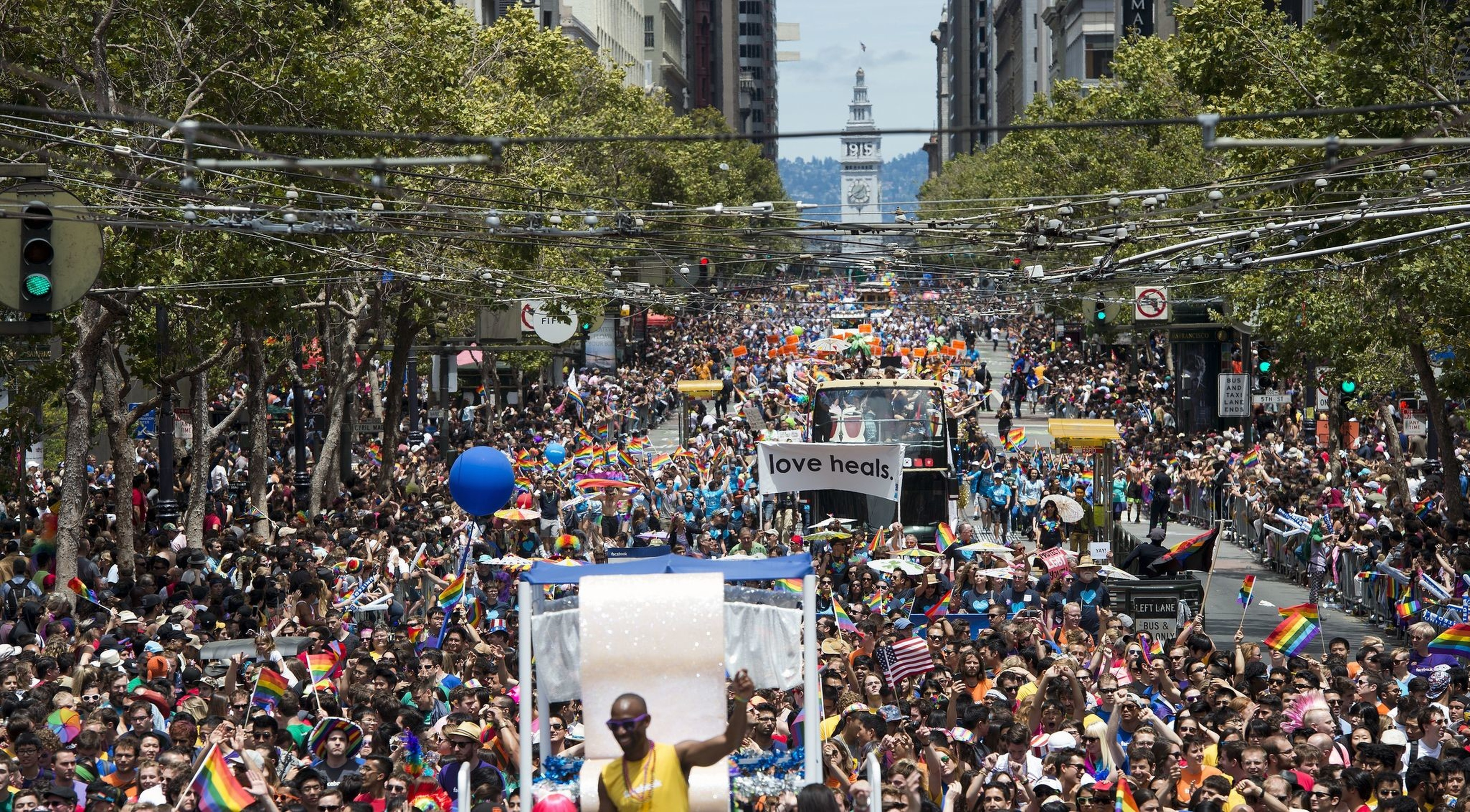 A massive crowd fills a city street during a Pride parade, waving rainbow flags and holding signs, with a clock tower visible in the background.