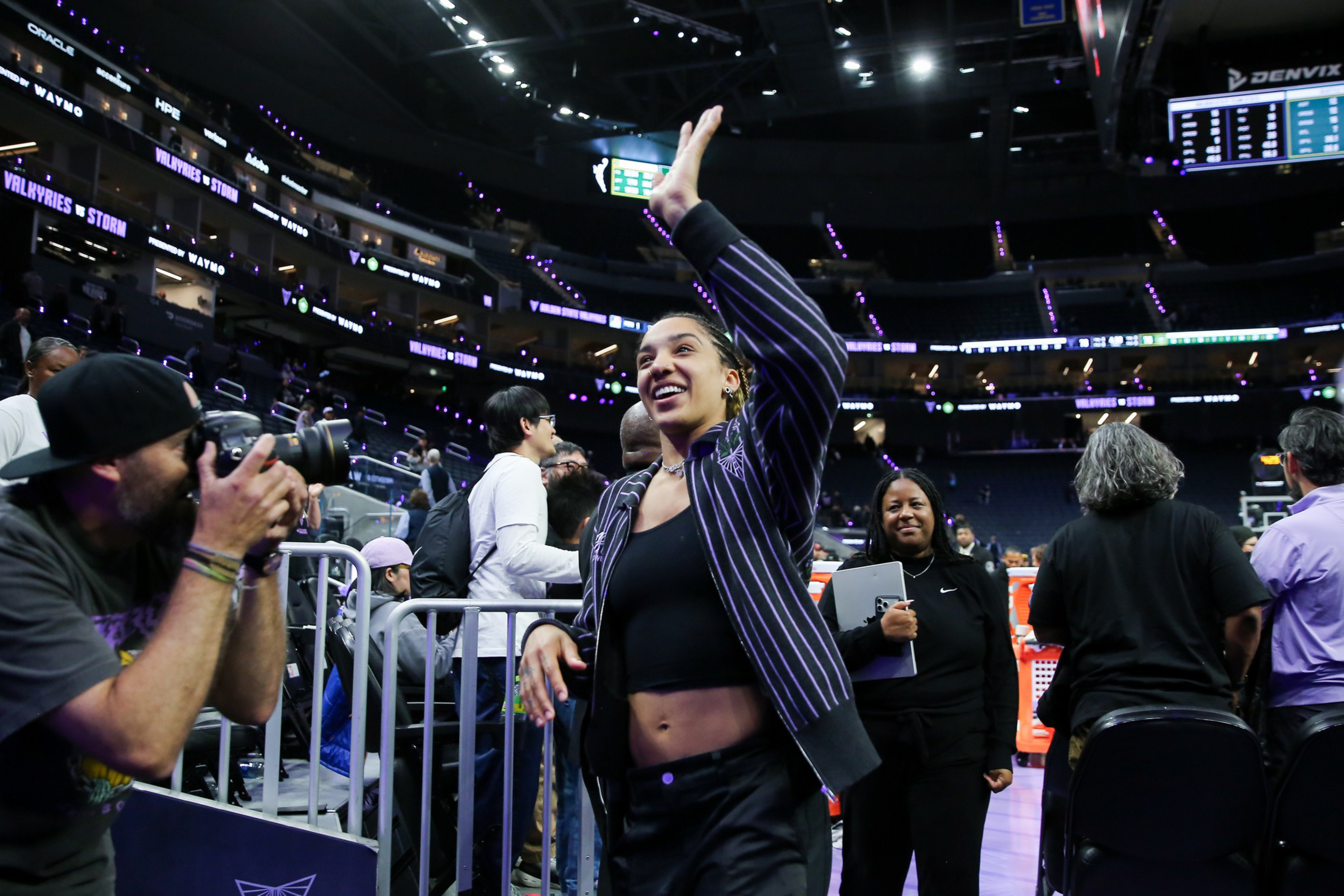 A woman in a striped jacket waves and smiles while a photographer takes her picture in a brightly lit indoor arena with spectators around.