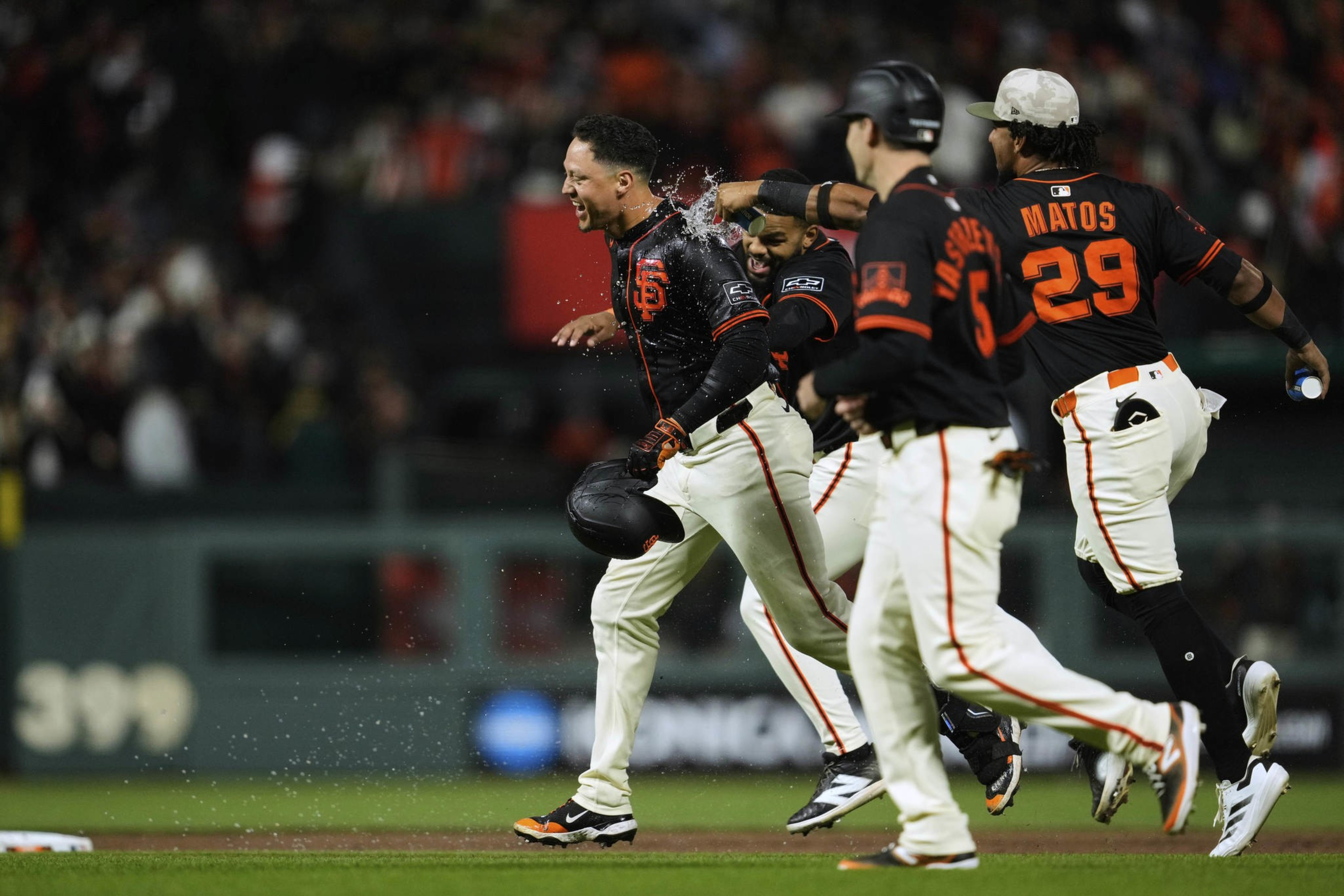 Players in black and orange baseball uniforms celebrate on a field, one throwing water over another who holds a helmet, with a cheering crowd in the background.