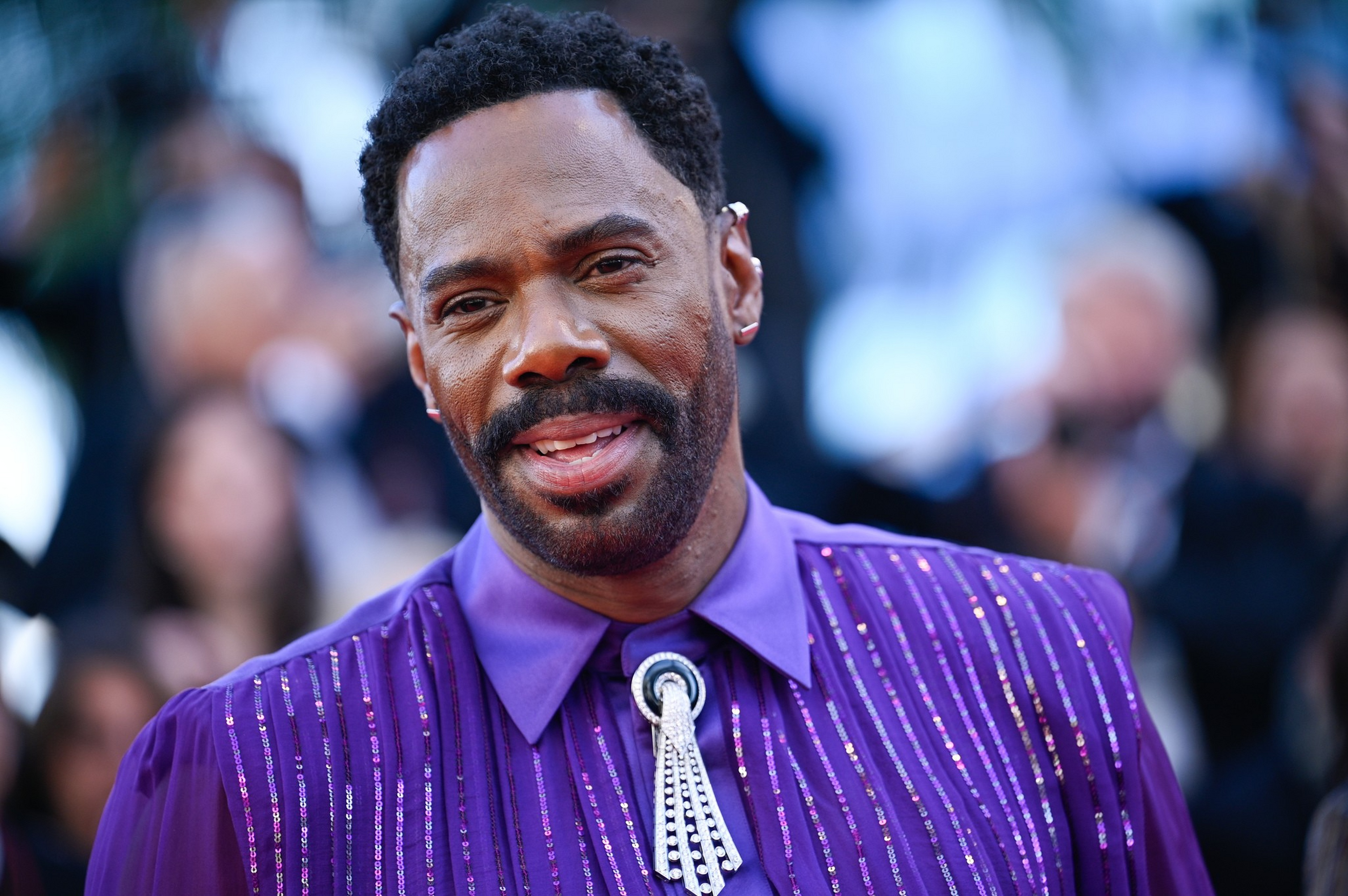 A man with short curly hair and a beard wears a shiny purple shirt with vertical stripes and a jeweled brooch, smiling slightly at the camera.