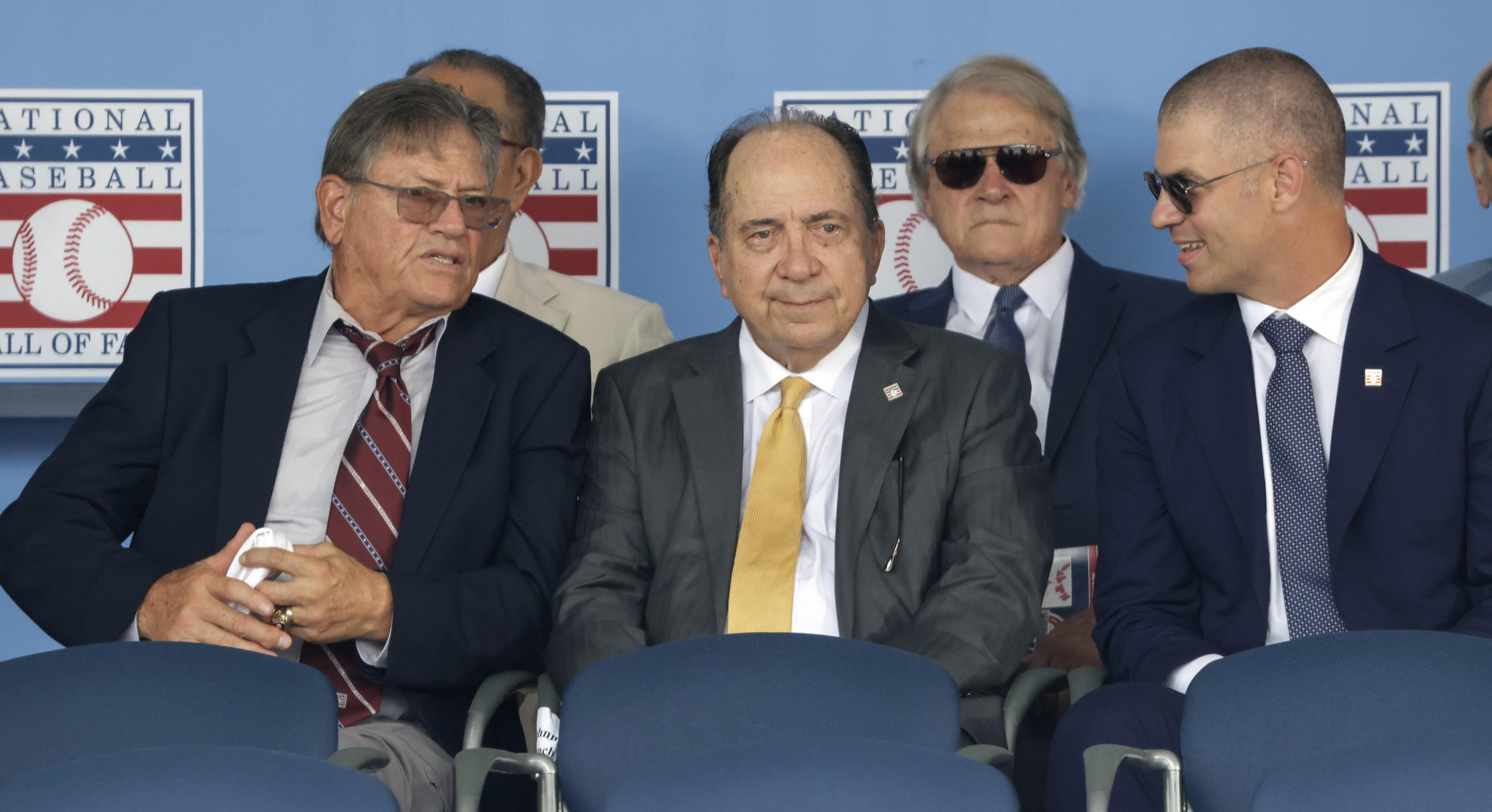 Five men in suits and sunglasses sit together, with a backdrop of the National Baseball Hall of Fame logo. They seem to be attending an event.