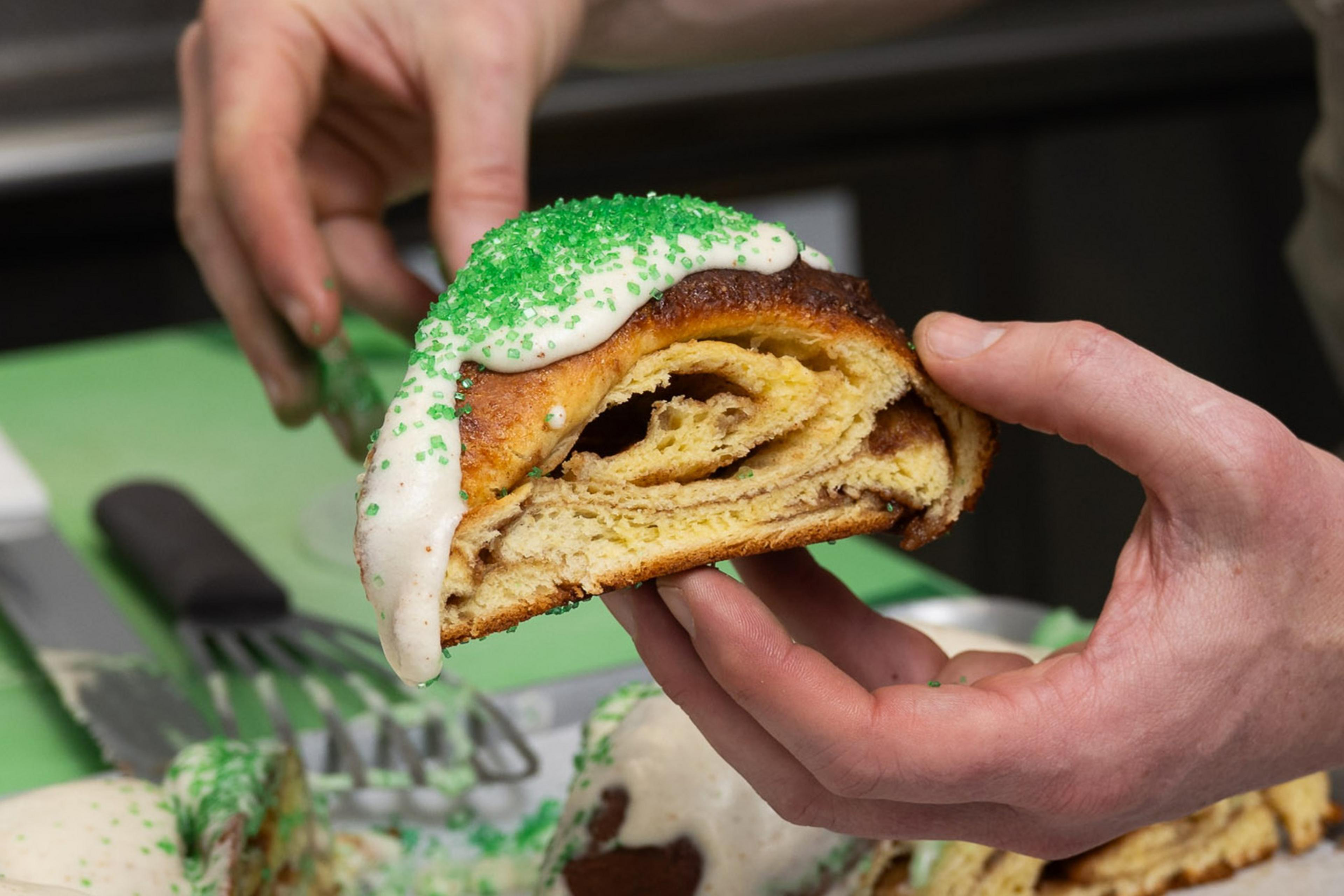 A person is holding a piece of pastry with swirled layers, topped with icing and green sprinkles. The background includes a spatula and a green surface.