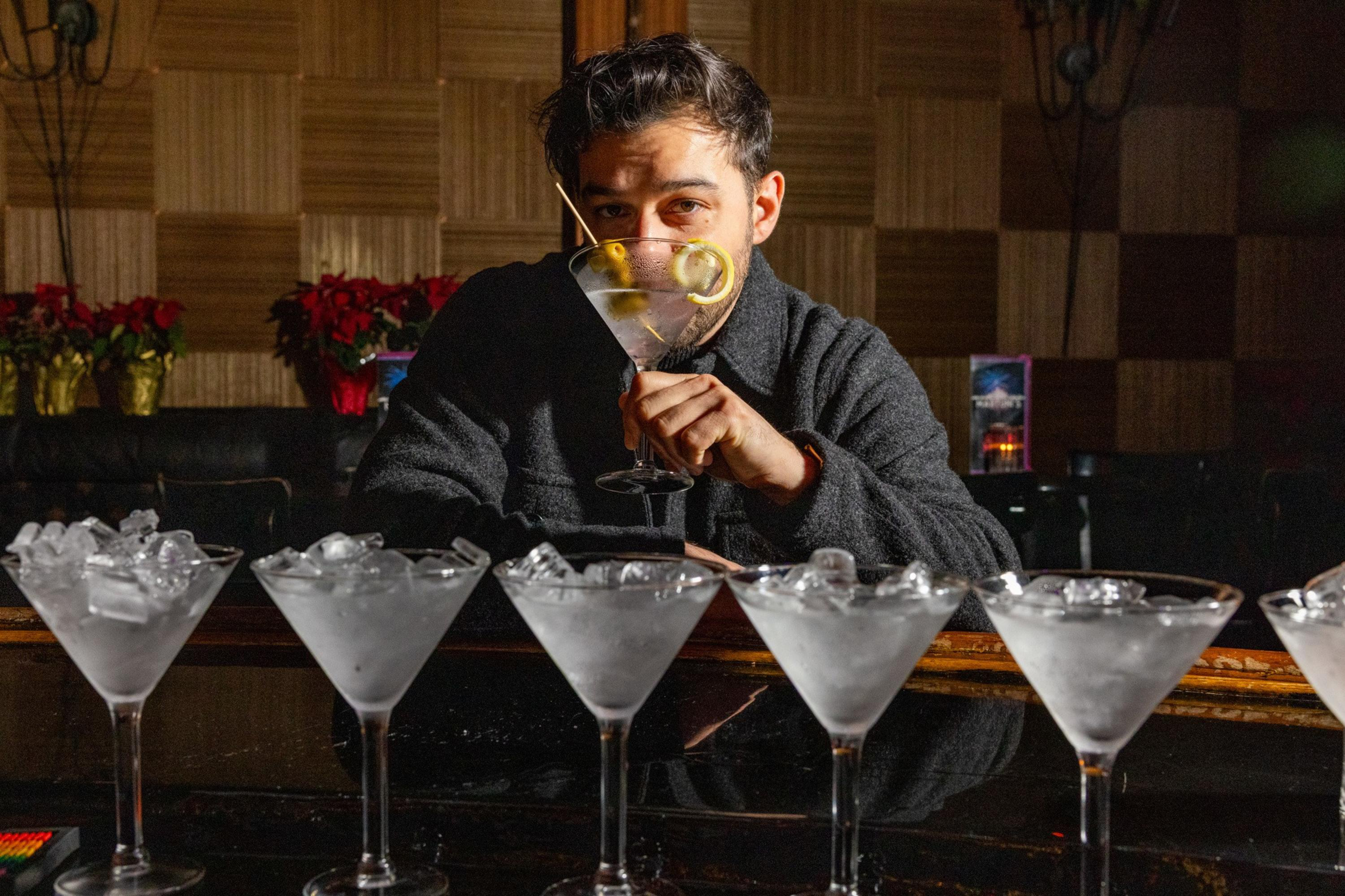 A man sits at a bar holding a martini glass with a lemon twist and olive, behind a row of six martini glasses filled with ice.