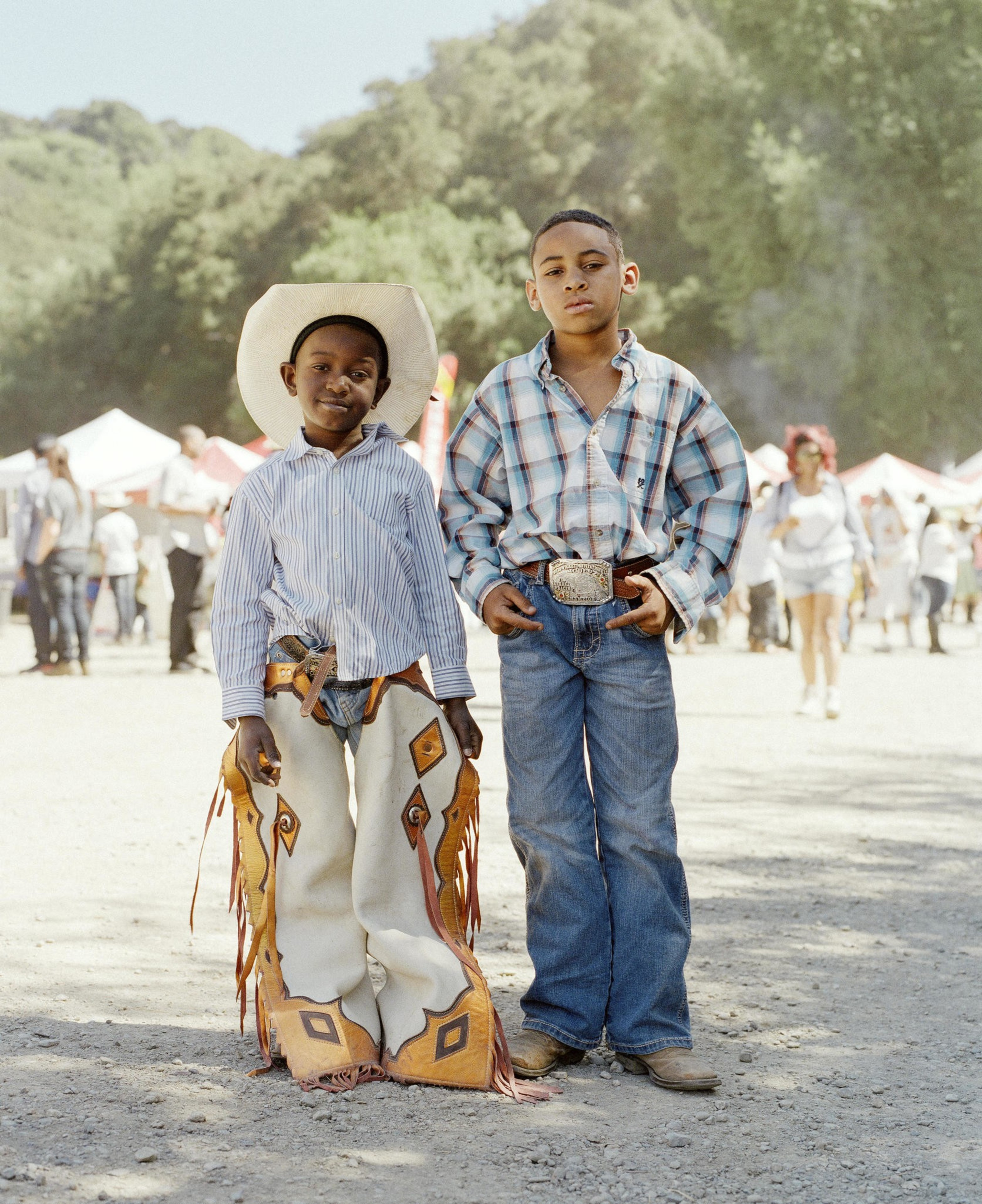 Two children stand outdoors, dressed in Western attire. One wears a cowboy hat and chaps, while the other has a plaid shirt and belt buckle. A festival is in the background.